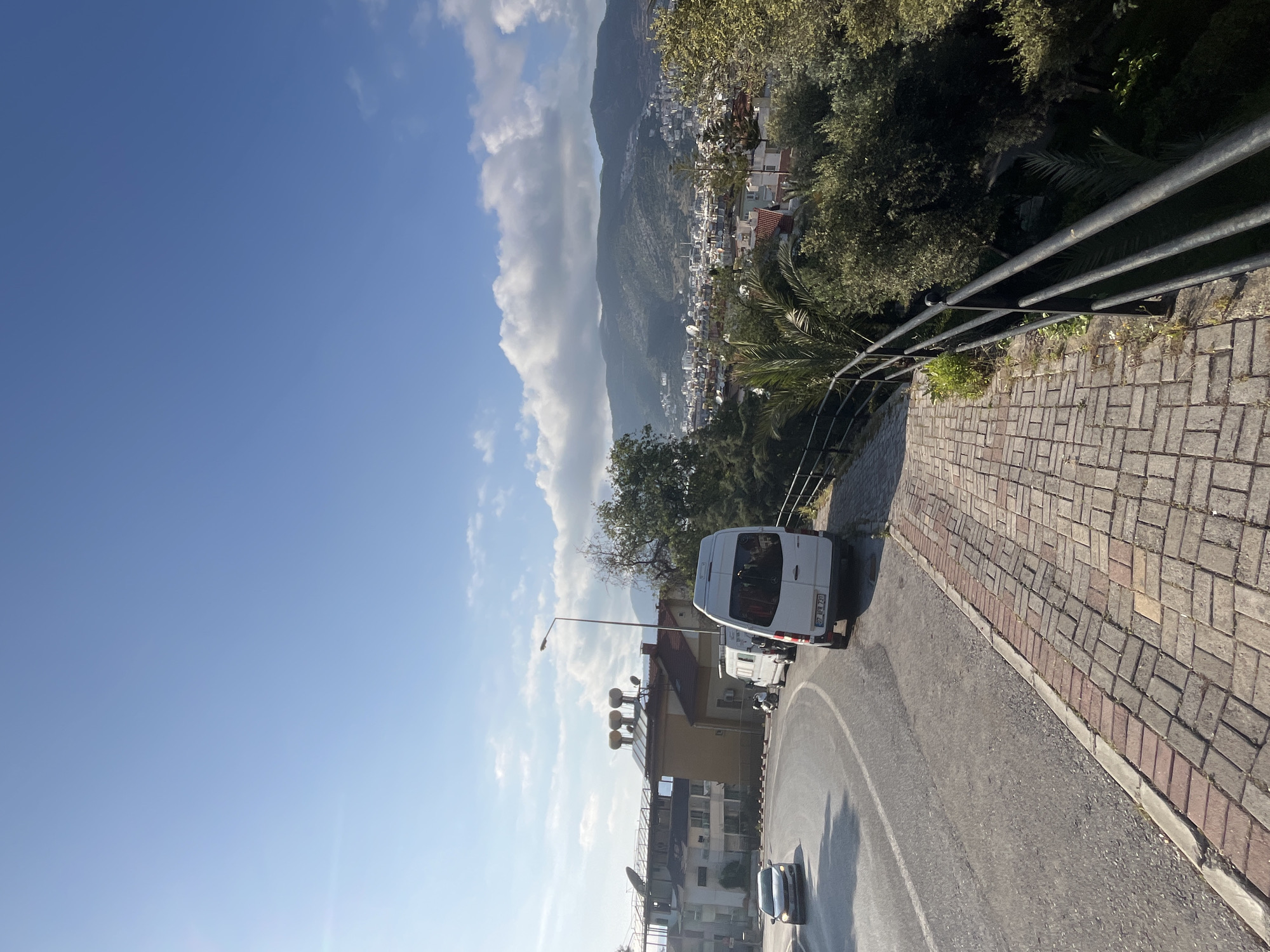 The image captures a vibrant, sunny landscape of Alanya, Turkey, from an elevated vantage point. The scene looks down upon a coastal town situated at the base of majestic, green-clad mountains under a bright blue sky.
In the foreground, a paved road curves from the left to the lower right of the frame. Alongside the road on the right, there is a patterned brick sidewalk bordered by a black metal railing. Lush green foliage, including palm trees, is visible along the roadside and in the valley below the railing.
On the road, a white minibus or van is prominently featured, facing away from the viewer. Its rear license plate, "38 P 8752", is clearly visible. Further along the curve of the road, a dark-colored car can also be seen. A lamppost stands near the white van.
The mid-ground reveals a sprawling urban area with numerous white or light-colored buildings, many featuring red-tiled roofs, extending towards the mountains. Satellite dishes and other rooftop structures are visible on some of these buildings. The background is dominated by large, imposing mountains, covered in dense greenery, stretching across the horizon beneath a sky dotted with scattered white clouds, particularly a larger cloud bank on the left.
The clear blue sky and strong shadows indicate a bright, sunny day, likely morning or afternoon. The overall impression is one of a picturesque and tranquil Mediterranean setting. No specific human activity or prominent event is depicted beyond the general presence of infrastructure and vehicles.