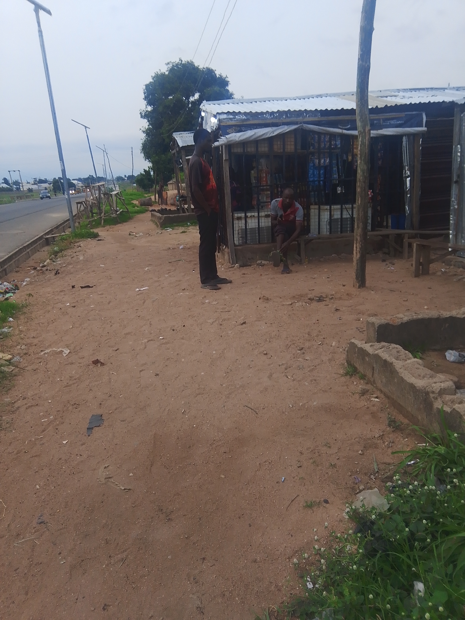 The image depicts a roadside scene in Jalingo, Nigeria, featuring two men standing near a makeshift shop. The shop, constructed with a corrugated metal roof and wooden supports, displays various goods behind a wire mesh. One man, wearing a red shirt and dark pants, stands in front of the shop, facing away from the camera. The second man, in a grey and red shirt and dark shorts, is seated on a wooden bench outside the shop, looking towards the ground. The foreground is dominated by sandy, uneven terrain with scattered debris, including plastic bags and wrappers. A paved road runs along the left side of the image, with a few vehicles visible in the distance. A tall, grey utility pole with several lights stands prominently on the left, and another wooden pole is situated near the shop. The background consists of green trees and an overcast sky. The overall environment suggests a relatively undeveloped, possibly peri-urban or rural setting within Jalingo.