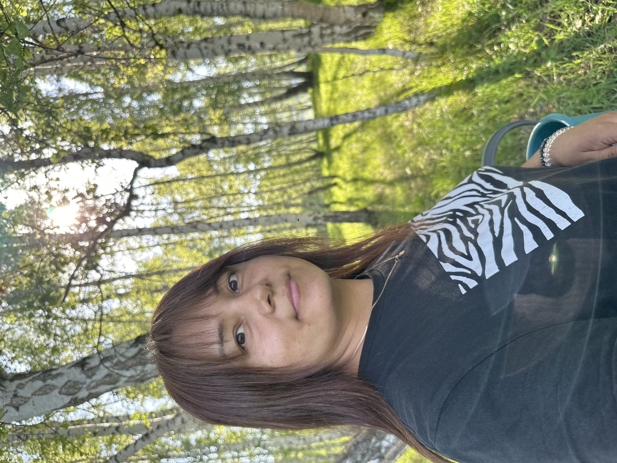 A woman is photographed outdoors in an area characterized by trees. The woman has brown hair with bangs, a tan complexion, and wears a black t-shirt with a white zebra-striped pattern. She is wearing multiple bracelets on her right wrist. The background consists of a row of birch trees with green leaves and grass on the ground. Bright sunlight is visible through the trees. Location: Unknown, Unknown.