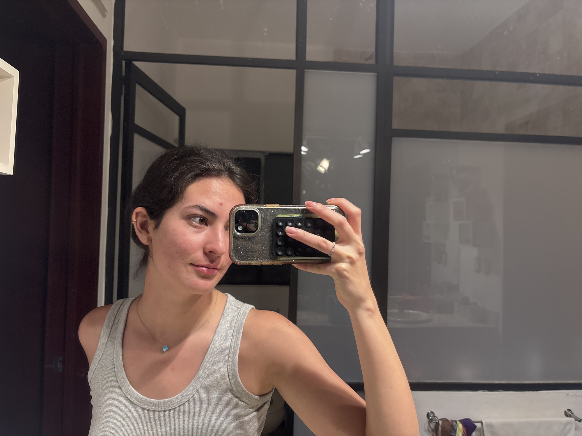 A woman takes a selfie in a bathroom. She is wearing a gray tank top and a delicate necklace with a blue stone. She is holding her phone with a glittery case and a pop socket. The bathroom has a dark wooden door on the left and a glass-paneled wall behind her. The location may be Puebla, Mexico, but this cannot be confirmed from the image alone.