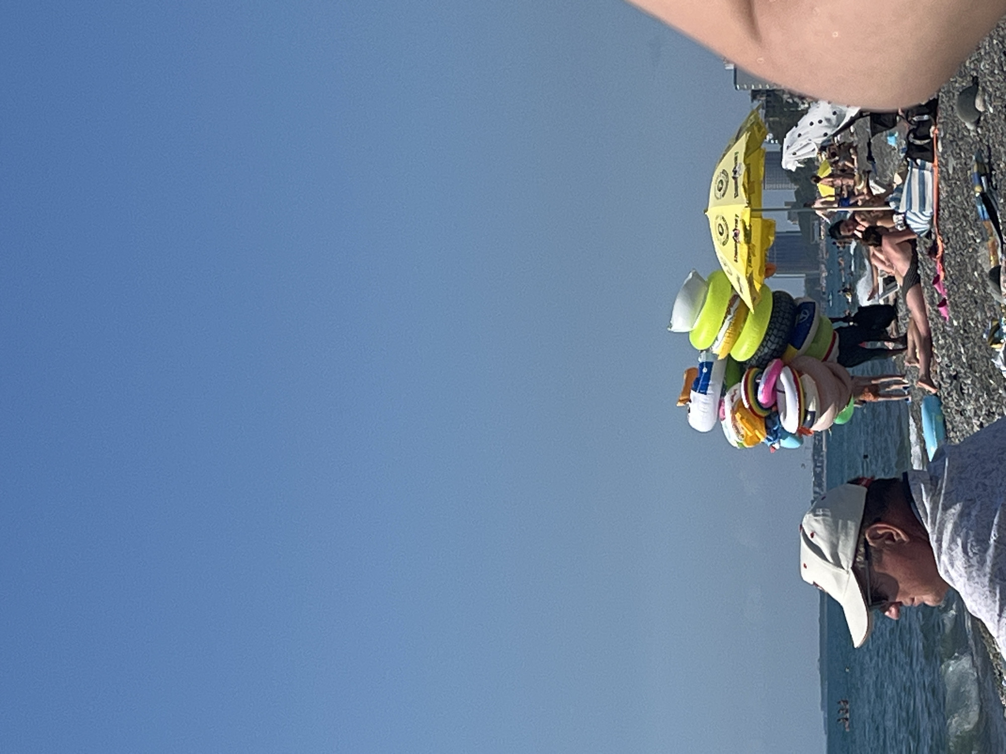 The image depicts a sunny day at the pebble beach in Kobuleti, Georgia. A man wearing a white baseball cap and sunglasses is in the foreground, looking out towards the sea. Behind him, a vendor is selling inflatable rings, stacked high on their shoulder. Several beachgoers are visible in the background, some relaxing under a yellow umbrella, while others are scattered across the beach and in the water. Buildings are visible in the distance, suggesting the presence of a town or city. The sea is calm, with small waves lapping the shore.