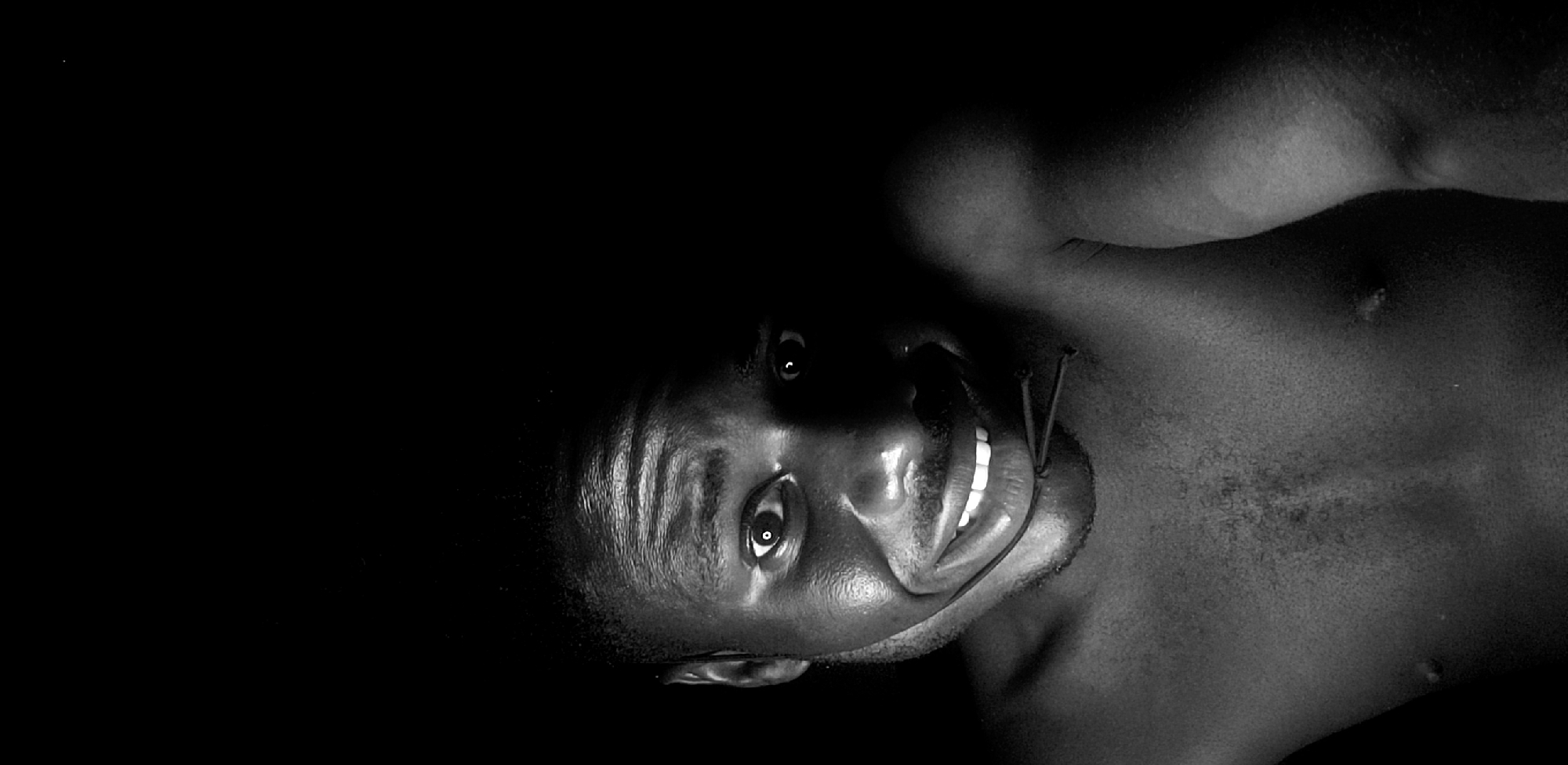 A low-angle, black and white photograph features a man from the chest up, turned to the right. He is smiling. A thin string wraps around his neck. The location is unknown and there is no mention of Narayi, Nigeria.