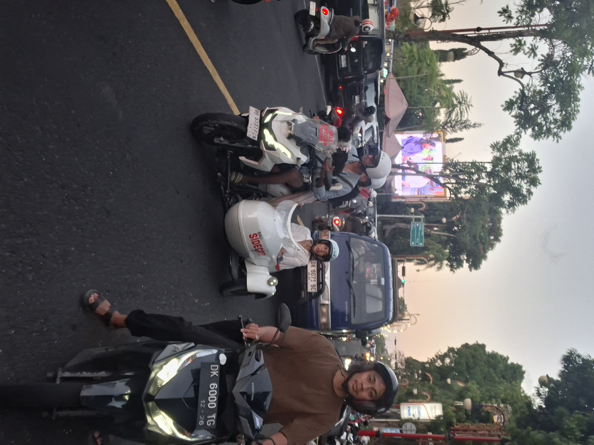 The image shows a street scene in Gianyar, Indonesia, likely during dusk. A man wearing a brown shirt and a black helmet is riding a black motorcycle with a license plate reading "DK 6000 TG". Beside him, a white motorcycle with a sidecar is being ridden by a man in a blue shirt and white helmet, carrying a child in the sidecar who is wearing a white shirt and blue helmet. The sidecar has "BALI SIDE CAR" written on it. In the background, various vehicles, including cars and other motorcycles, are present in traffic. Trees line the street, and a large digital billboard displaying people is visible in the distance. The sky has a hazy, twilight appearance.