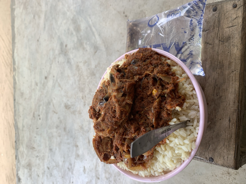 The scene depicts a meal set on a wooden surface in Ikot Ete Ekpo, Nigeria. A light pink-rimmed bowl, positioned centrally, contains white rice topped with a generous portion of reddish-brown stew. The stew features visible pieces of meat or fish and sliced onions. A silver-colored metal spoon is vertically inserted into the food within the bowl. To the right of the bowl, a clear plastic sachet of drinking water, partially filled, rests on the same wooden surface. Blue markings and a droplet logo are visible on the sachet. The wooden surface itself is light brown, appears aged, and is composed of at least two planks, secured by visible nail heads. The background consists of a light grey, textured ground surface, possibly concrete or packed earth, illuminated by natural light. No individuals are present in the image.