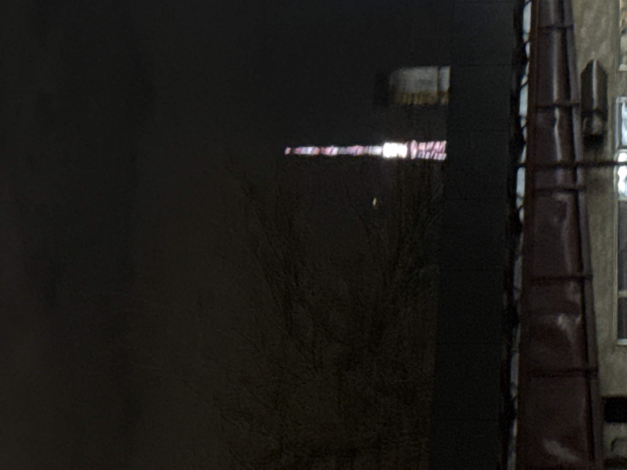This low-light image captures an urban night scene, likely in Yerevan, Armenia. The left and central portions are dominated by a vast, dark, reflective surface, possibly a window or a shadowed building facade. Within this dark expanse, faint outlines of bare tree branches or bushes are subtly visible in the lower-center.
A striking horizontal band of bright, elongated light segments spans the middle of the reflective surface. These segments vary in color from intense white to soft pink and red, appearing as blurry reflections of illuminated signs or windows from a distant structure. While the forms within this light band resemble abstract patterns, they suggest segments of unreadable illuminated text or symbols due to the distortion and low resolution. Above this, a fainter, rectangular reflection is noticeable in the upper-central dark area.
To the right, a section of an adjacent building is partially visible. It features a prominent brownish-red vertical structure, possibly a downspout or a decorative architectural element, running along its side. Further up, details of an upper floor can be discerned, including a faint window outline and what appears to be an external unit, possibly an air conditioner.
The overall scene is characterized by deep shadows and the prominent glow of artificial lights reflected against the dark surfaces, indicating it was captured at night or in very low light conditions. No specific activity or individuals are present in the static scene.