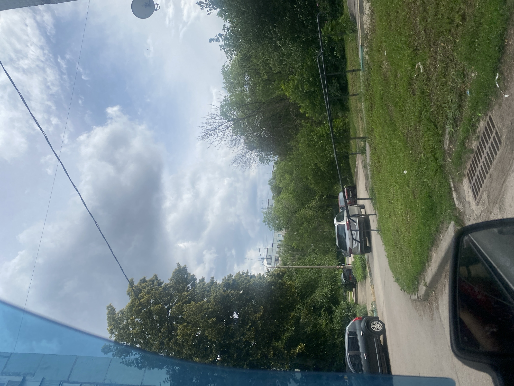 The image depicts a view from a vehicle, looking out at a residential street in Samara, Russia. The foreground shows the reflection of a person in the car's side mirror. The street is paved, with a curb and a grassy area to the right, featuring a storm drain. Several cars are parked or driving on the street. In the middle ground, there are trees and greenery, with a light-colored building in the background. The sky is cloudy.