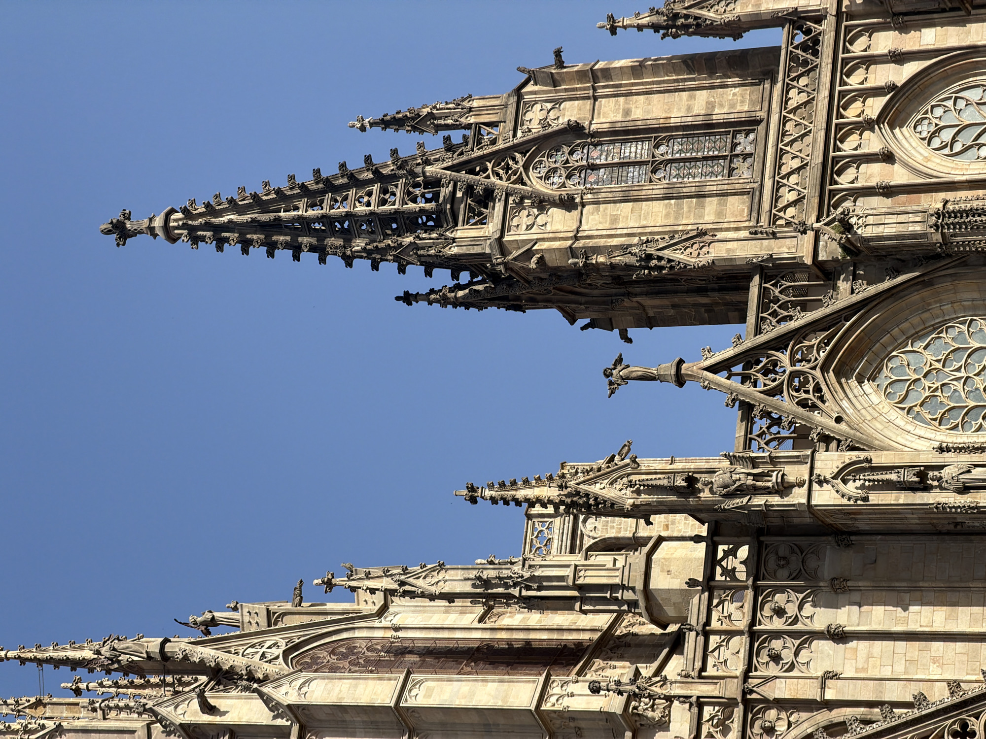 The image shows a close-up, low-angle view of the facade of the Barcelona Cathedral (Catedral de Barcelona) in Barcelona, Spain. The architectural style is Gothic, characterized by intricate stonework, pointed arches, spires, and statues. The cathedral is constructed from weathered stone, with visible details of its ornate carvings, gargoyles, and stained-glass windows. The sky is a clear, deep blue, providing a stark contrast to the ancient architecture. The sunlight casts shadows that highlight the three-dimensional texture of the building. There are no people visible in the image. The focus is solely on the architectural grandeur of the cathedral.