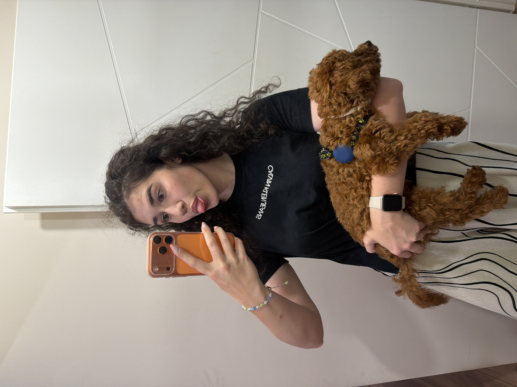 A young woman with long, curly dark hair stands in what appears to be a residential hallway or entryway, possibly in Sofia, Bulgaria. She is holding an orange smartphone in her right hand, taking a selfie, and playfully sticking out her tongue towards the camera. She is wearing a black t-shirt with white lettering that reads "CALVIN KLEIN JEANS" and a long, light-colored skirt with vertical black stripes. On her left wrist, she wears a beaded bracelet with blue, green, and white beads. She is holding a small, curly-haired brown dog in her left arm. The dog is wearing a blue collar with yellow paw print accents. The background consists of neutral-toned walls and a white door or closet panel with geometric lines.