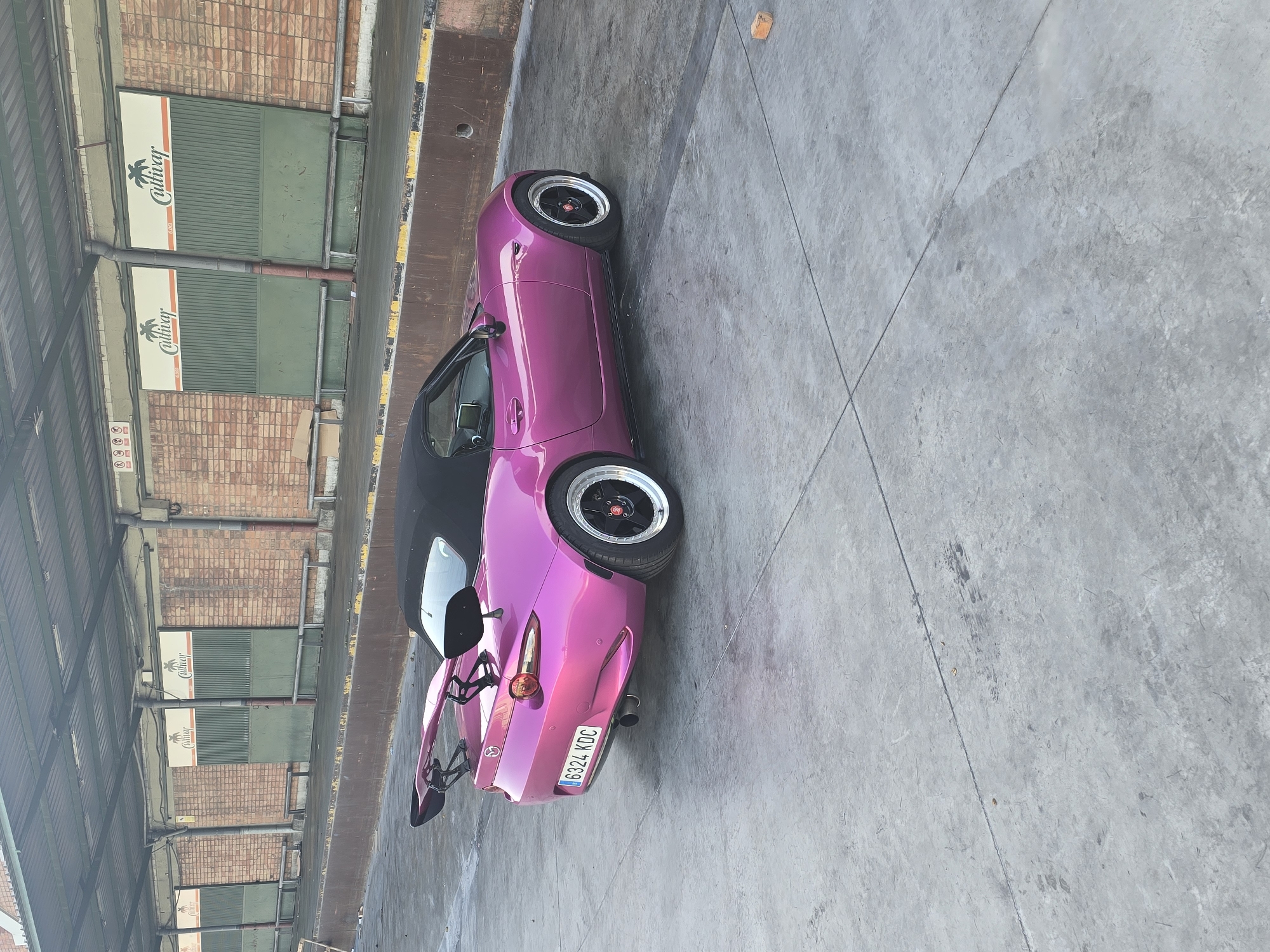A bright pink two-door convertible sports car is parked outdoors on a concrete surface. The car, featuring a black soft top, a prominent black rear spoiler/wing, and custom black and silver multi-spoke wheels, is oriented sideways to the viewer, facing slightly left. A Spanish license plate, "6324 KDC", is visible on the rear of the vehicle.
The setting appears to be an industrial or commercial area, possibly a loading dock or warehouse facility. In the background, there is a building with a brick facade and several large, dark green garage-style doors. Above these doors, white signs are affixed, prominently displaying the word "Cultivay" with a palm tree logo, and the word "BOX" underneath. A metal roof structure extends over the area where the car is parked, providing shade. The lighting indicates it is daytime with clear, sunny weather. No people are visible in the scene.