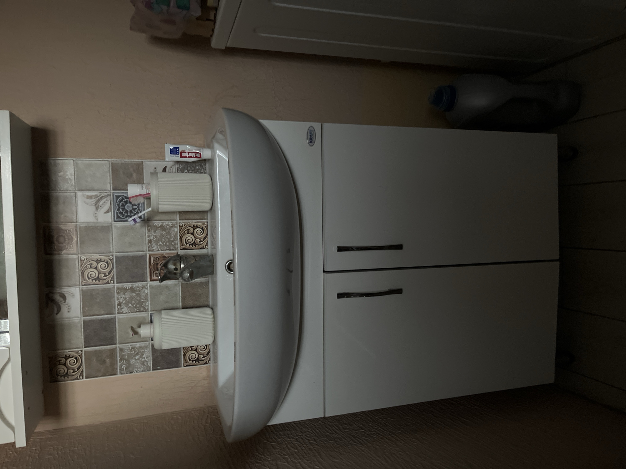 A medium shot shows a white sink vanity with two cabinet doors and two handles against a wall with tiled backsplash and beige textured wallpaper. A white dispenser and a faucet are on the sink. To the right of the faucet, a white cup holds a toothbrush and a tube of toothpaste. Next to the cup is a tube of "Dr. MORGAN" toothpaste. A grey laundry machine is to the right of the vanity. A large grey bottle with a blue cap sits on the floor to the right of the vanity.