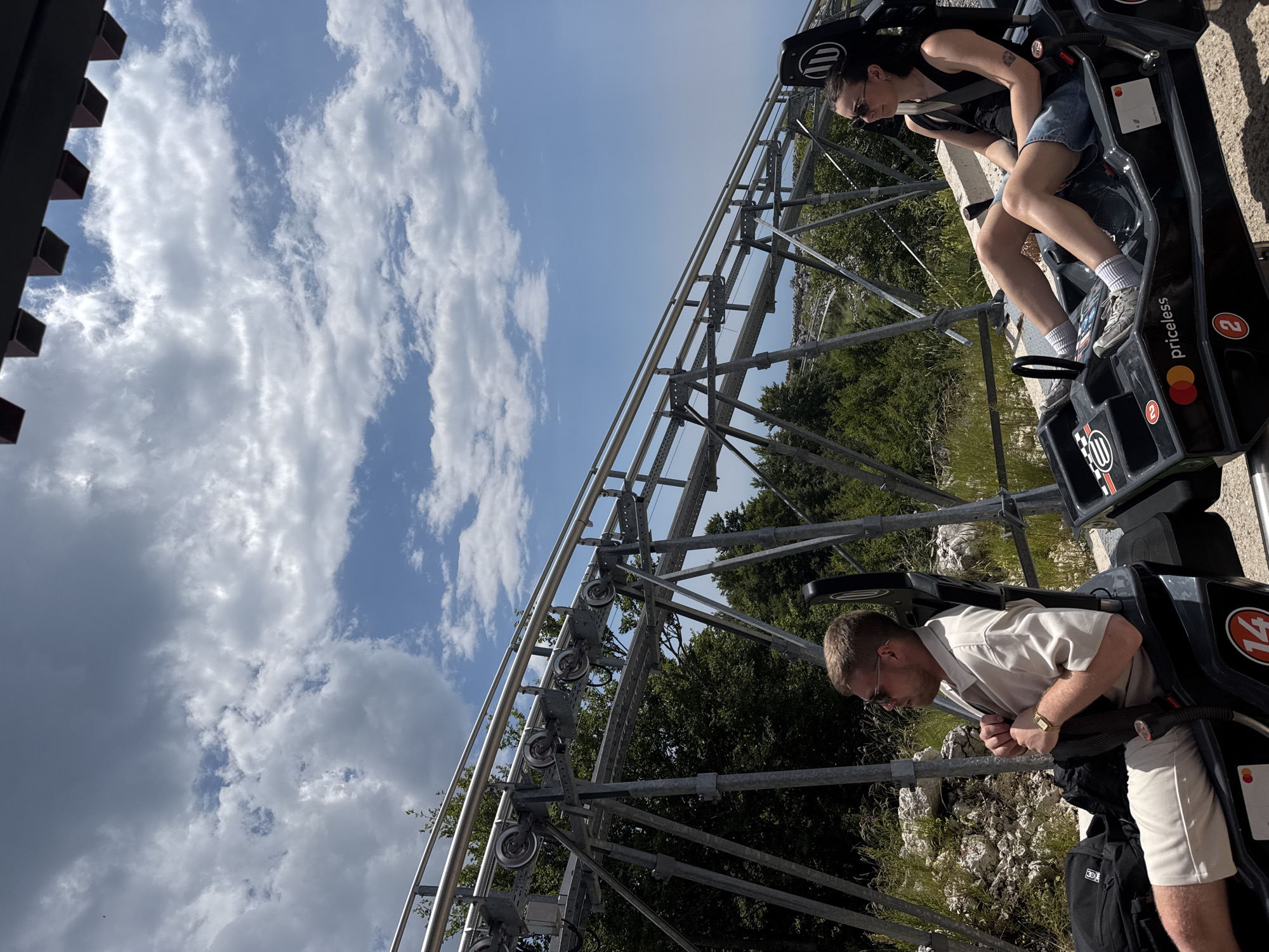 This media file depicts two individuals on a roller coaster or similar gravity-powered ride, likely at an outdoor attraction. The perspective is from a slightly elevated angle looking down at the riders and the track. The sky is a bright blue with scattered white clouds, suggesting it is daytime and the weather is fair. Lush green trees and rocky terrain are visible in the background, hinting at a natural or mountainous location.
The person in the foreground, a man, is wearing a light-colored collared shirt and beige shorts. He is seated in the ride's cart, leaning forward, and appears to be gripping the safety harness. His expression is difficult to discern, but his posture suggests anticipation or excitement. He is wearing sunglasses and a wristwatch.
In the background, another individual, a woman, is seated in a cart ahead of the man. She is wearing a black tank top, denim shorts, and sunglasses. Her head is turned slightly, and she also appears to be looking forward.
The ride's structure is prominently featured, with metal tracks and supports visible. The carts are dark-colored with some branding. Text visible on the carts includes "priceless," a Mastercard logo, and the number "2" within a red circle. Another cart shows the number "14" in a similar red circle. The overall scene conveys an atmosphere of outdoor recreation and thrill-seeking. The location context of Žanjev Do, Montenegro, suggests this might be a specific tourist attraction in that region.