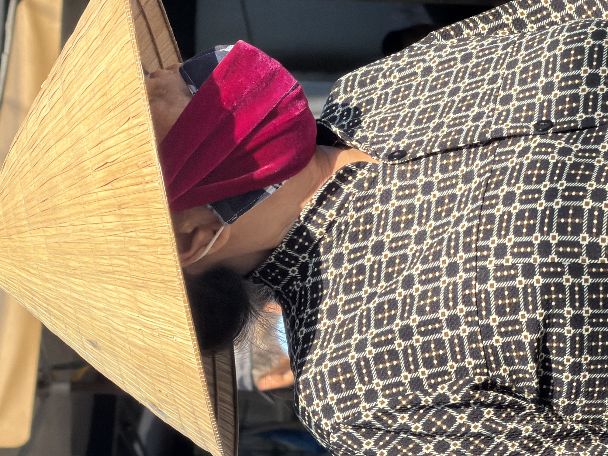 The image shows a person wearing a conical hat, a face mask, and a patterned shirt. The person's face is partially obscured by the hat and mask, but their ear is visible. The shirt has a geometric pattern with dark brown, white, and small brown dots. The conical hat is made of woven material and casts a shadow on the person's face. The face mask is a deep pink or magenta color, with a darker patterned fabric peeking out from underneath. The background is blurred and suggests an outdoor market setting, possibly in Nha Trang, Vietnam, given the conical hat, which is a traditional Vietnamese headwear.
