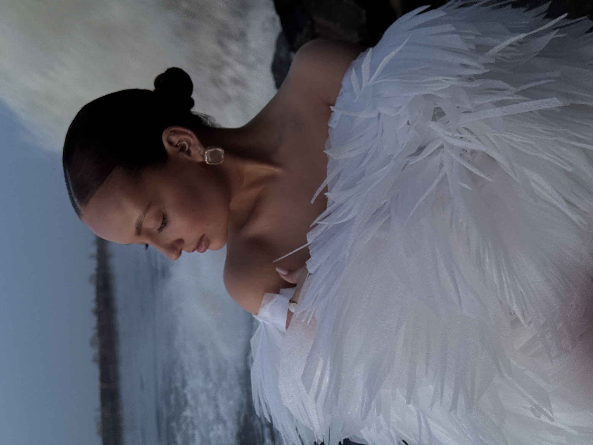 A woman is depicted in a photograph taken outdoors, in Chaikovsky, Russia. She has dark hair styled in a bun, and is wearing square stud earrings. The woman is wearing a garment or costume of white, feathery material covering one shoulder and a portion of her torso. The background features a body of water with a waterfall effect, suggesting a riverside or dam location. The overall composition of the image is taken at an angle and the environment suggests a natural yet controlled landscape.