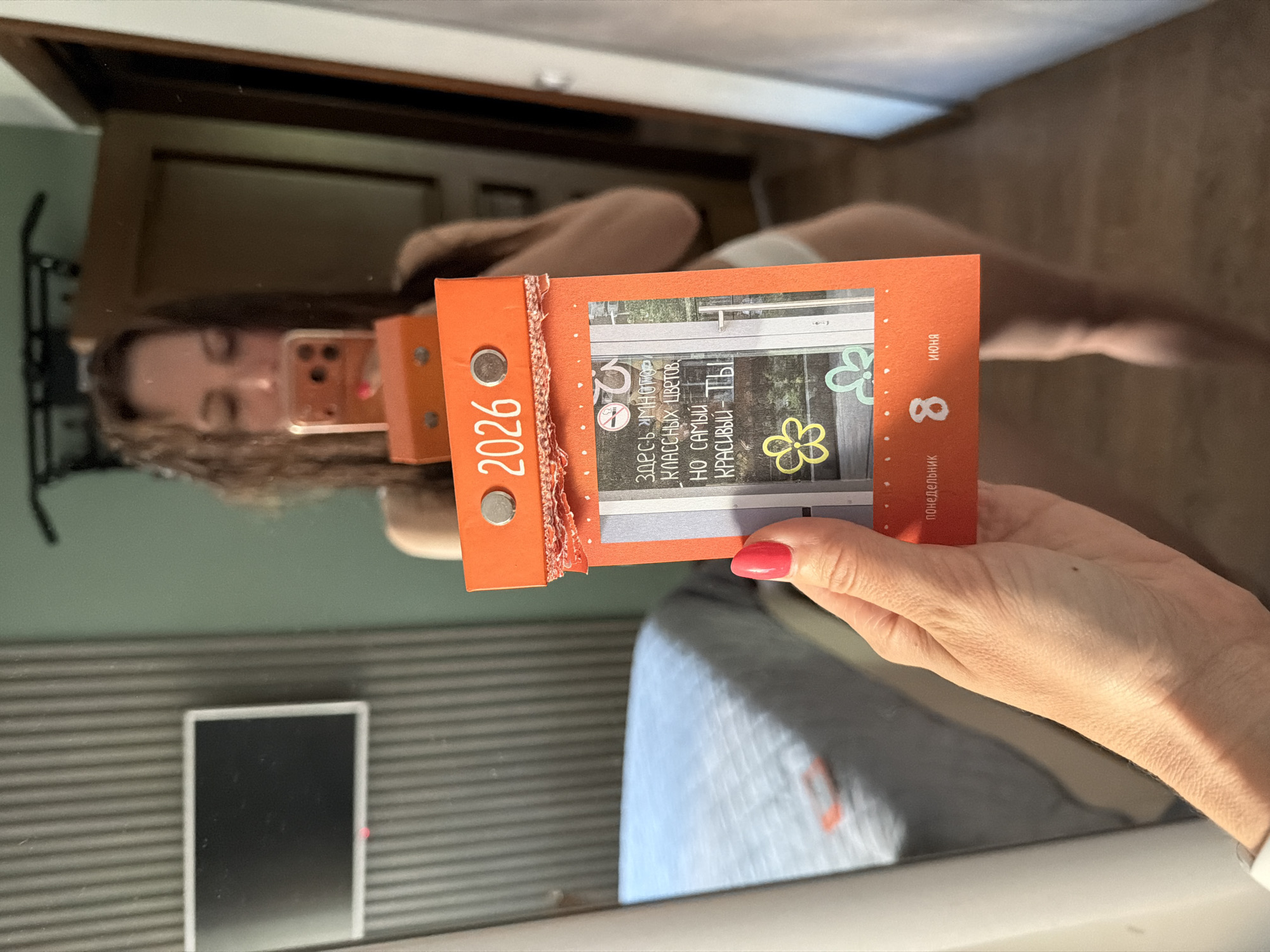 A woman is taking a selfie in a mirror. She is holding an orange calendar that says "2026" and has the date "8 ИЮНЯ" (June 8th) on it. The calendar also has a message written in Russian that translates to "There are many beautiful flowers here, but you are the most beautiful." The background shows a bedroom with a bed and a television on the wall. The scene appears to be indoors, likely in a home. There is no indication of the city of Birobidzhan, Russia, in the image.