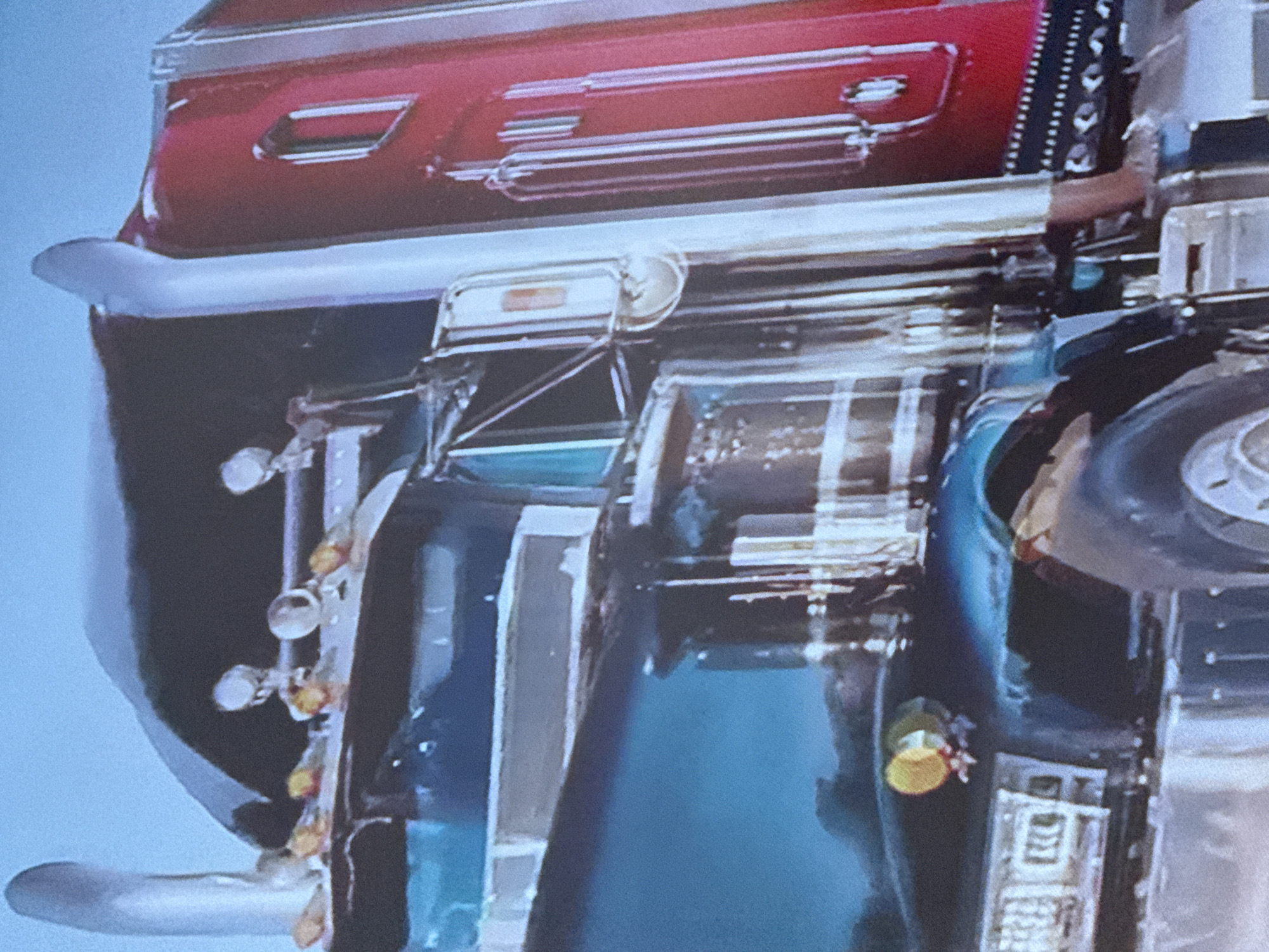 The image depicts the underside of a vehicle, possibly a truck, photographed from a low angle. The vehicle features a red rear section with chrome detailing, including taillights and trim. The lower portion of the vehicle is predominantly blue, with various mechanical components visible, such as a wheel, suspension elements, and a bumper. The image's orientation is rotated, which may be the reason for the unusual perspective. The environment is blurry and indistinct, making it impossible to provide any details about the location, especially about the city of Ivanteyevka, Russia.