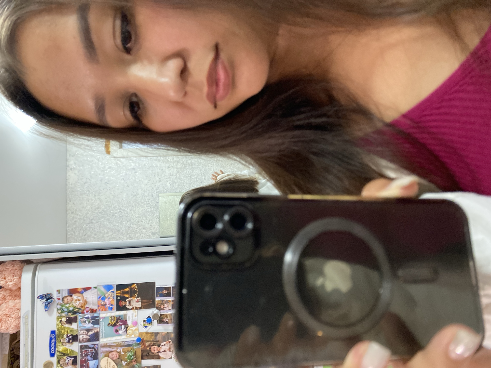 In a medium close-up, a young woman with long, dark hair takes a selfie in what appears to be a kitchen, possibly in the city of Ufa, Russia. She is wearing a bright magenta top. Her phone, with a black case and an Apple logo visible on the back, is held up, obscuring her face from the chest up. In the background, a refrigerator is adorned with numerous photographs. The lighting suggests it is daytime, with a bright overhead light source.