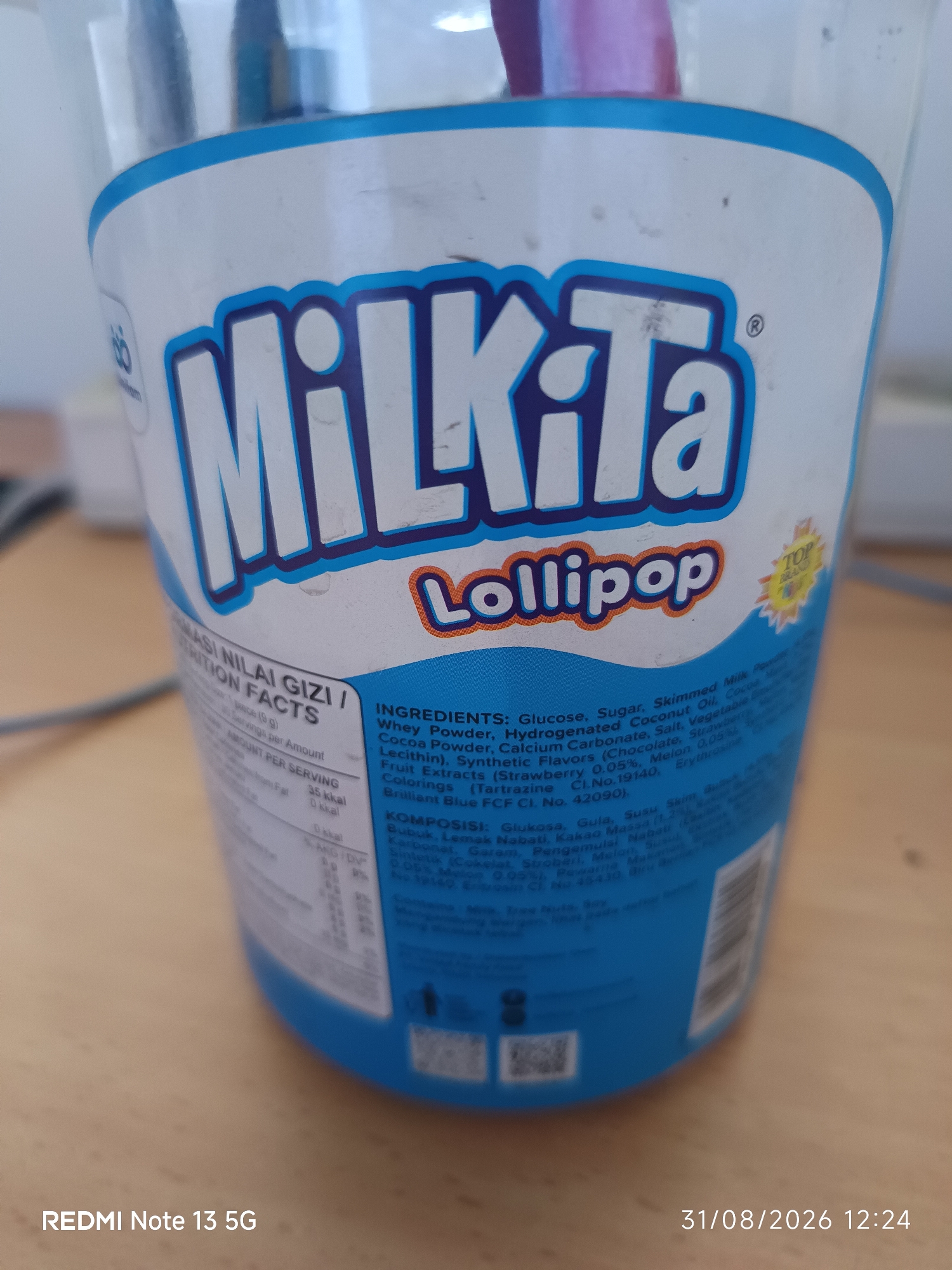 The image shows a close-up of a plastic container of "Milkita Lollipop" candies. The container is predominantly white with blue accents and text. The brand name "Milkita" is prominently displayed in large, blue, outlined letters, with "Lollipop" written below it in a smaller, orange font. The nutritional information and ingredients list are visible on the side of the container in both English and what appears to be Indonesian. Some of the ingredients listed in English include Glucose, Sugar, Skimmed Milk Powder, and various flavors. The container appears to be placed on a wooden surface. The bottom right corner of the image displays "REDMI Note 13 5G" and the date "31/08/2026 12:24," suggesting the image was taken with this phone model on that date and time. There is no visible indication of the location being the City of Medan, Indonesia.