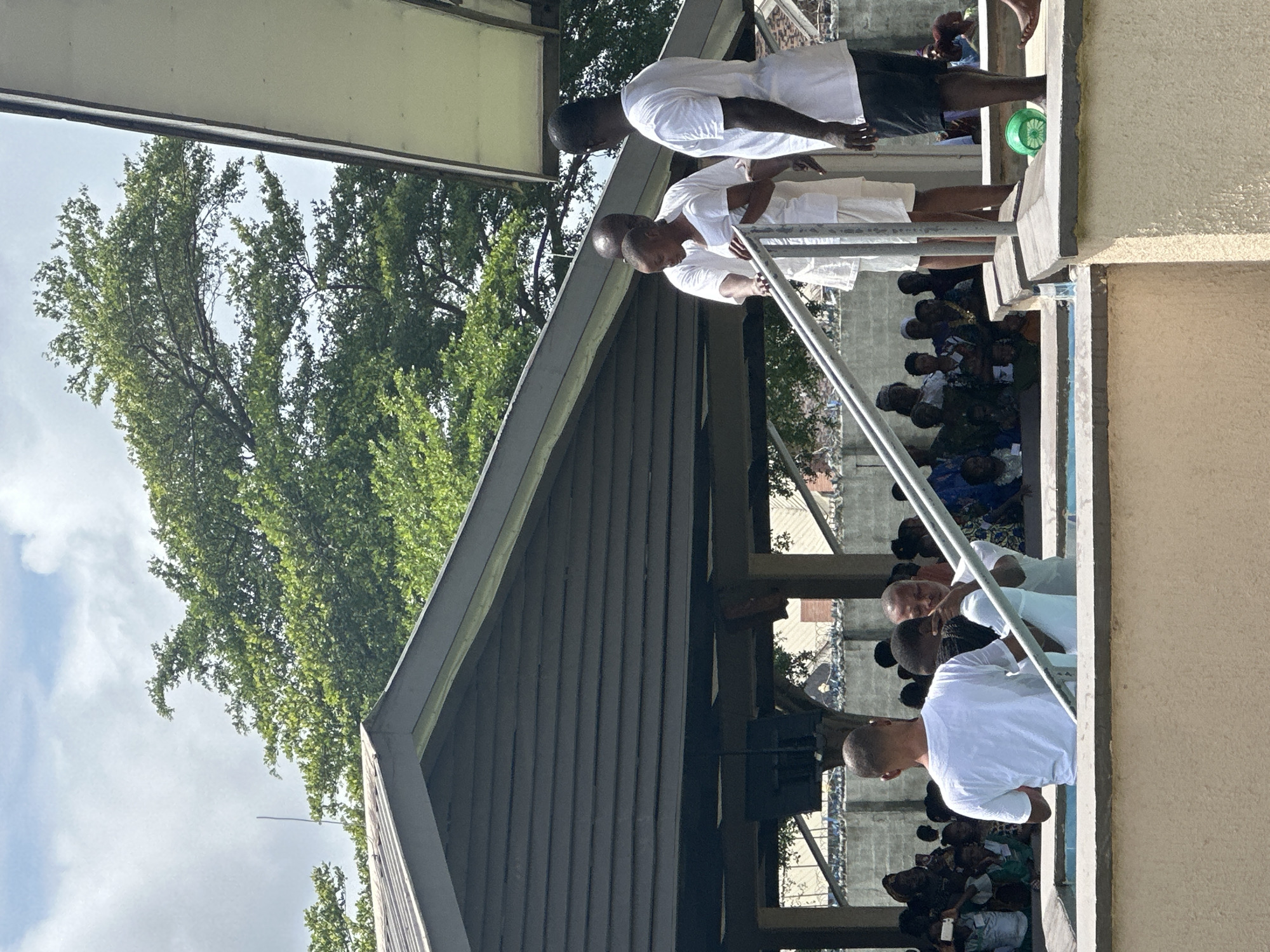 The image depicts an outdoor baptismal service in Itoga, Nigeria. In the foreground, three men in white t-shirts are positioned around a baptismal pool. One man, seen from the back, is partially submerged in the water, with another man on his left and a third man on his right, who is holding a metal railing. Behind them, a crowd of people, dressed in various attire, is gathered, observing the ceremony. The background features a covered structure with a corrugated roof and wooden beams, with lush green trees and a cloudy sky overhead. The overall scene suggests a religious gathering within a community setting.