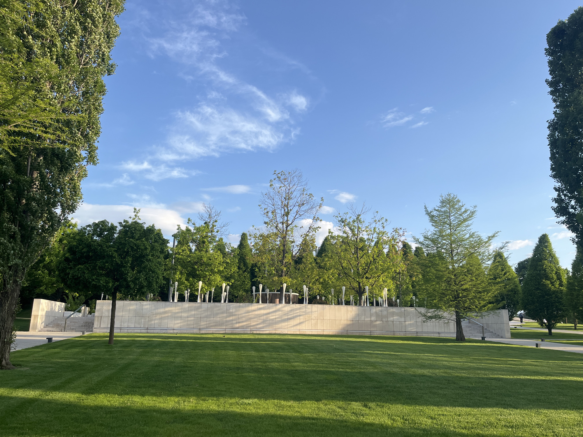 The image depicts a park scene on a sunny day in Krasnodar, Russia. A large, meticulously manicured green lawn dominates the foreground, displaying patterns from recent mowing. Behind the lawn is a long, low beige wall. Emerging from behind the wall are numerous white, pole-like sculptures, adding an artistic element to the landscape. Beyond the wall and sculptures, a variety of trees, including evergreens and deciduous types, create a dense backdrop. To the left and right sides of the frame, tall, mature trees partially obscure the view, framing the scene. The sky is blue with scattered wispy clouds, suggesting clear weather conditions. The overall impression is of a well-maintained, peaceful public space.