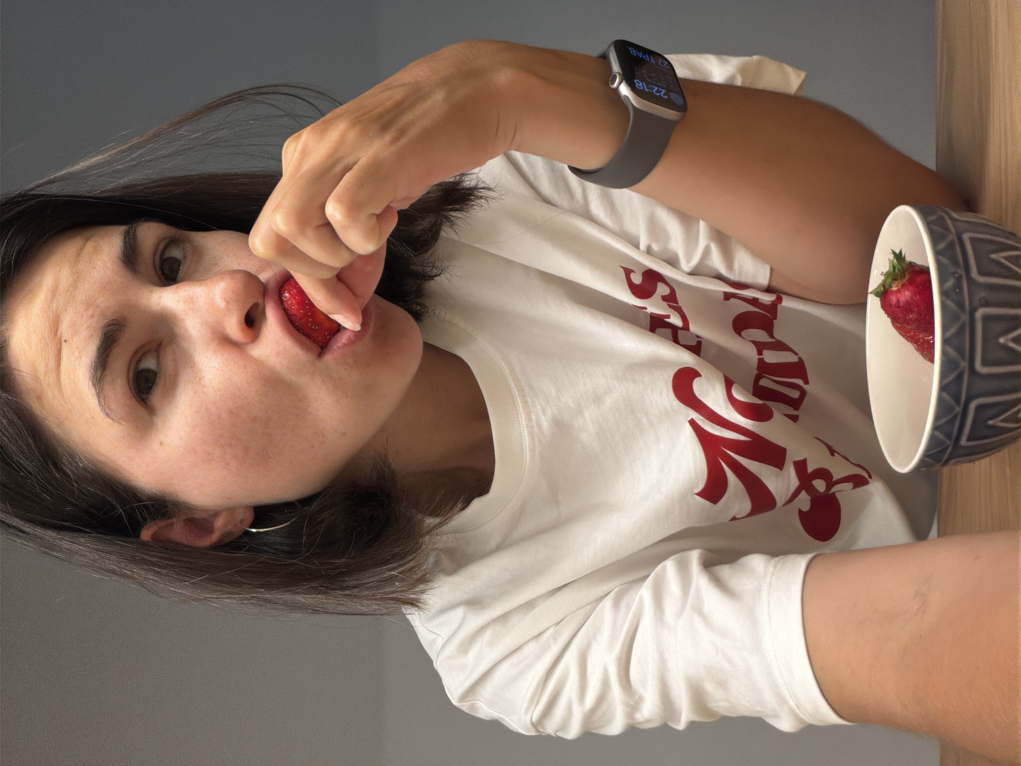 A woman is indoors, positioned at an angle to the camera. She is eating a strawberry, holding it with her fingers as it touches her lips. She wears a white T-shirt with red lettering. A watch is on her left wrist. In the upper right corner, a bowl contains another strawberry. The room has gray and wood tones. No specific location in Budva, Montenegro can be determined from this image.