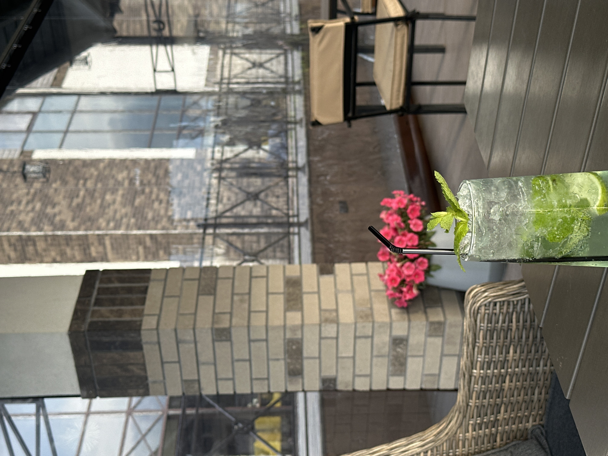 The image depicts a close-up of a glass of mojito cocktail on a table, with a wicker chair and a vase of pink flowers in the background. A brick column and a building with large windows are also visible. The scene appears to be an outdoor seating area, possibly a cafe or restaurant, with a fence and more chairs visible in the distance. The lighting suggests it is daytime, with soft natural light.