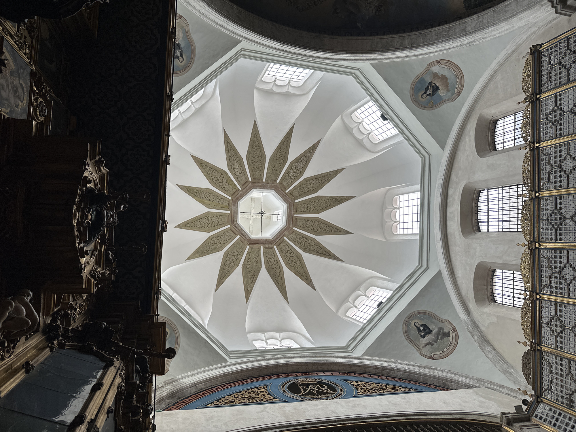 The image depicts the ceiling of a building in Mexico City, Mexico. The ceiling is a dome with an ornate, star-shaped pattern in the center. Around the star are several windows that let in natural light. Near the edges of the dome are portraits, likely of religious figures. The dome is intricately decorated with moldings and ornamentation, including patterned panels with symbols. The overall impression is of an elaborate and historical architectural space. The image is taken from a low angle, looking up at the ceiling.