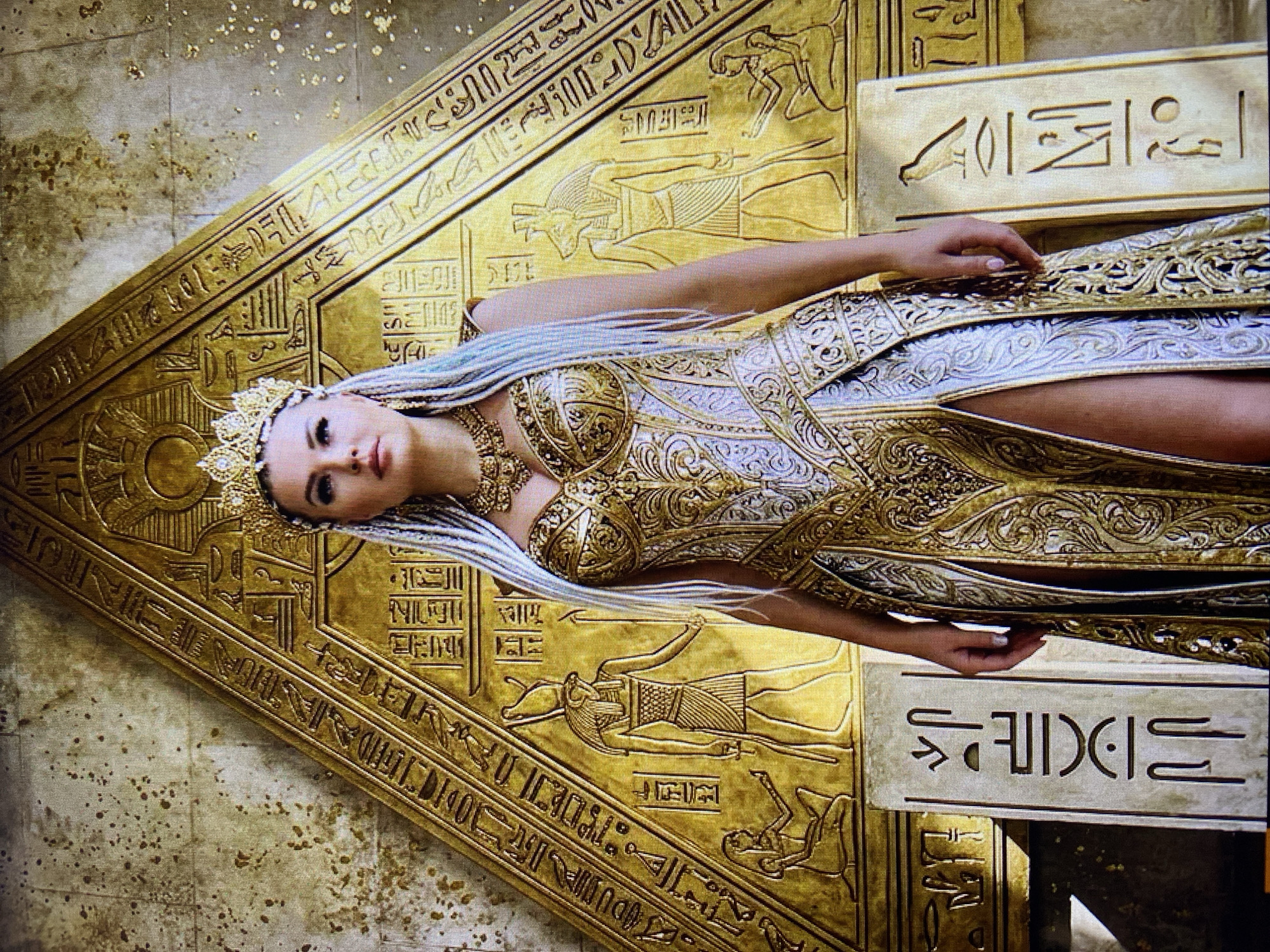 A woman in a bejeweled golden and white gown and a crown stands in front of a large, gold pyramid-shaped relief adorned with hieroglyphs and Egyptian deities. The woman has long, light-colored hair with blue streaks and is wearing heavy gold jewelry. The relief features figures of gods with animal heads and human forms, as well as smaller figures. Two stone tablets with carved hieroglyphs stand on either side of the relief at the woman's feet. The background is a textured, stone-like wall with speckled gold accents. The scene evokes an ancient Egyptian aesthetic. The image is not located in Stavropol, Russia.