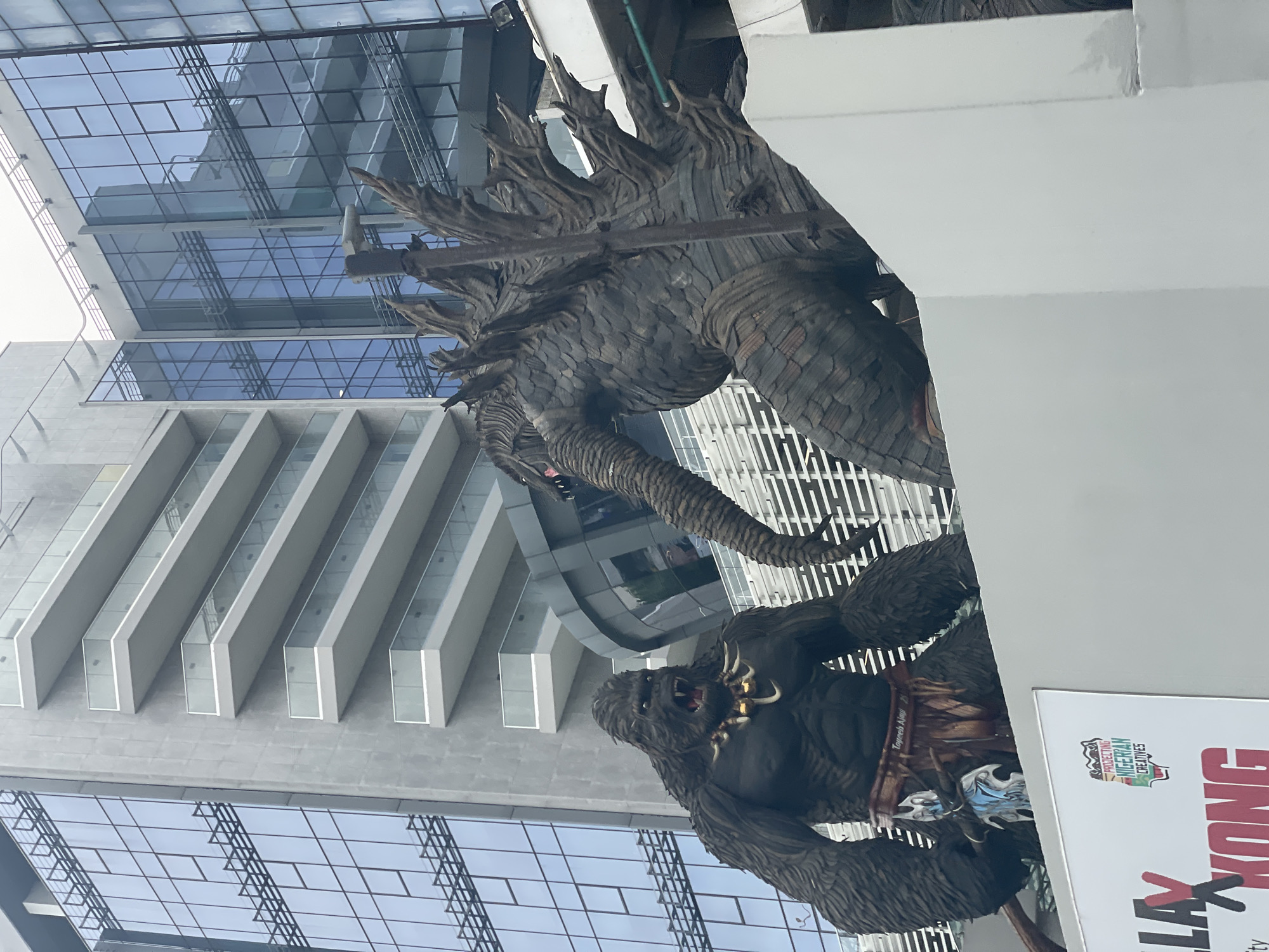 A large outdoor sculpture of Godzilla and King Kong is displayed in front of modern buildings in Itirin, Nigeria. The buildings feature glass and concrete construction. The sculptures are brown and appear to be made of sculpted metal. A sign is next to the sculptures with the words "Godzilla Kong" and "Protecting Nigerian Creatives."