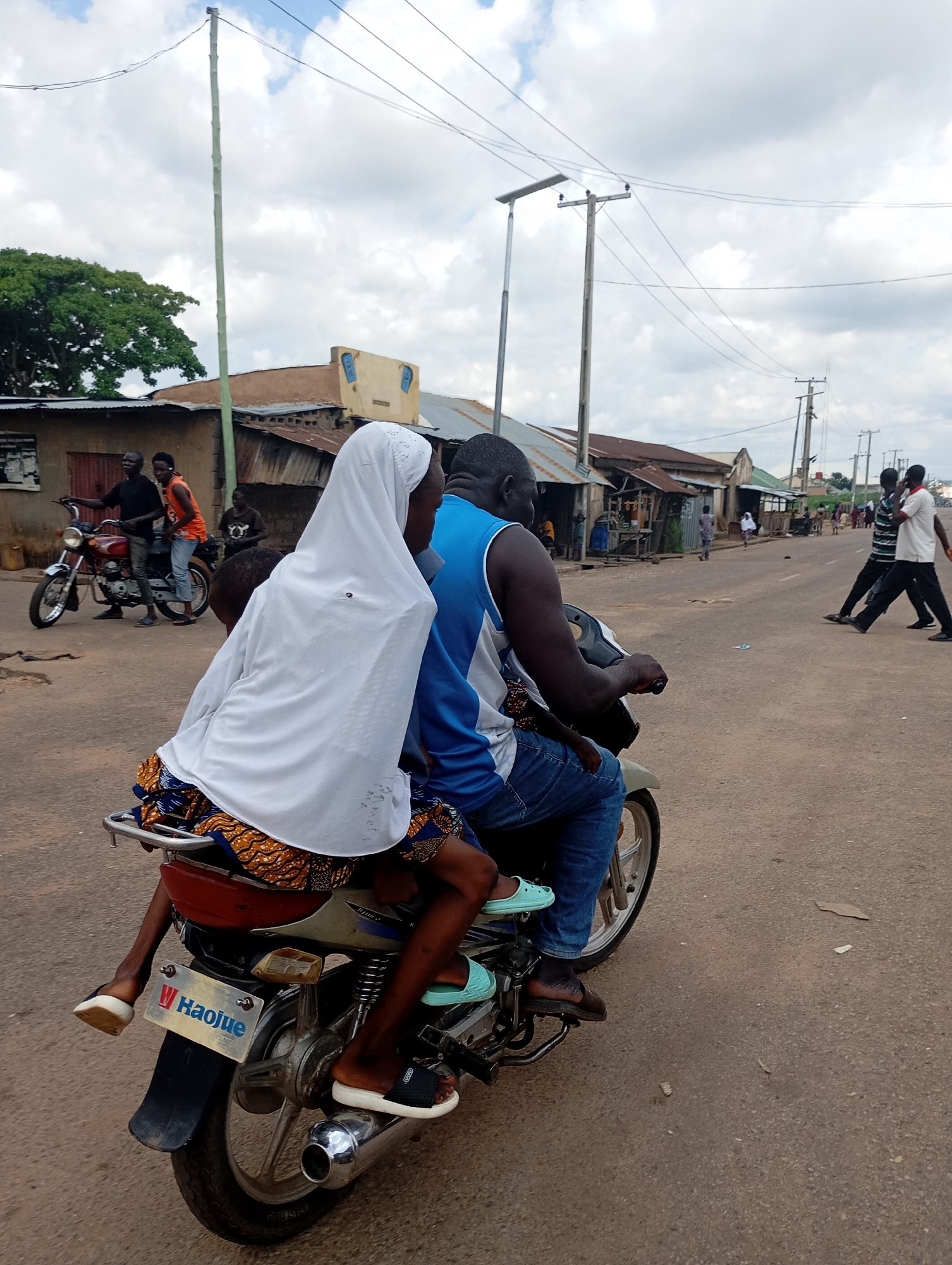 A medium-shot photograph captures two individuals on a motorcycle on a paved road in Shendam, Nigeria. The motorcycle is moving away from the viewer, with the rider and passenger facing away from the camera. The rider, a dark-skinned man, wears a blue sleeveless t-shirt and blue jeans. The passenger, a woman, is dressed in a white hijab and a patterned skirt. Her legs are dangling on the side of the motorcycle. The motorcycle has a visible license plate that reads "HaoJue". In the background, several buildings with corrugated iron roofs line the street, suggesting a commercial or residential area. A few other people are visible in the distance, some walking and others on a motorcycle. Utility poles and power lines are prominent in the sky, indicating the presence of infrastructure. The overall scene depicts everyday life on a road in a Nigerian town.