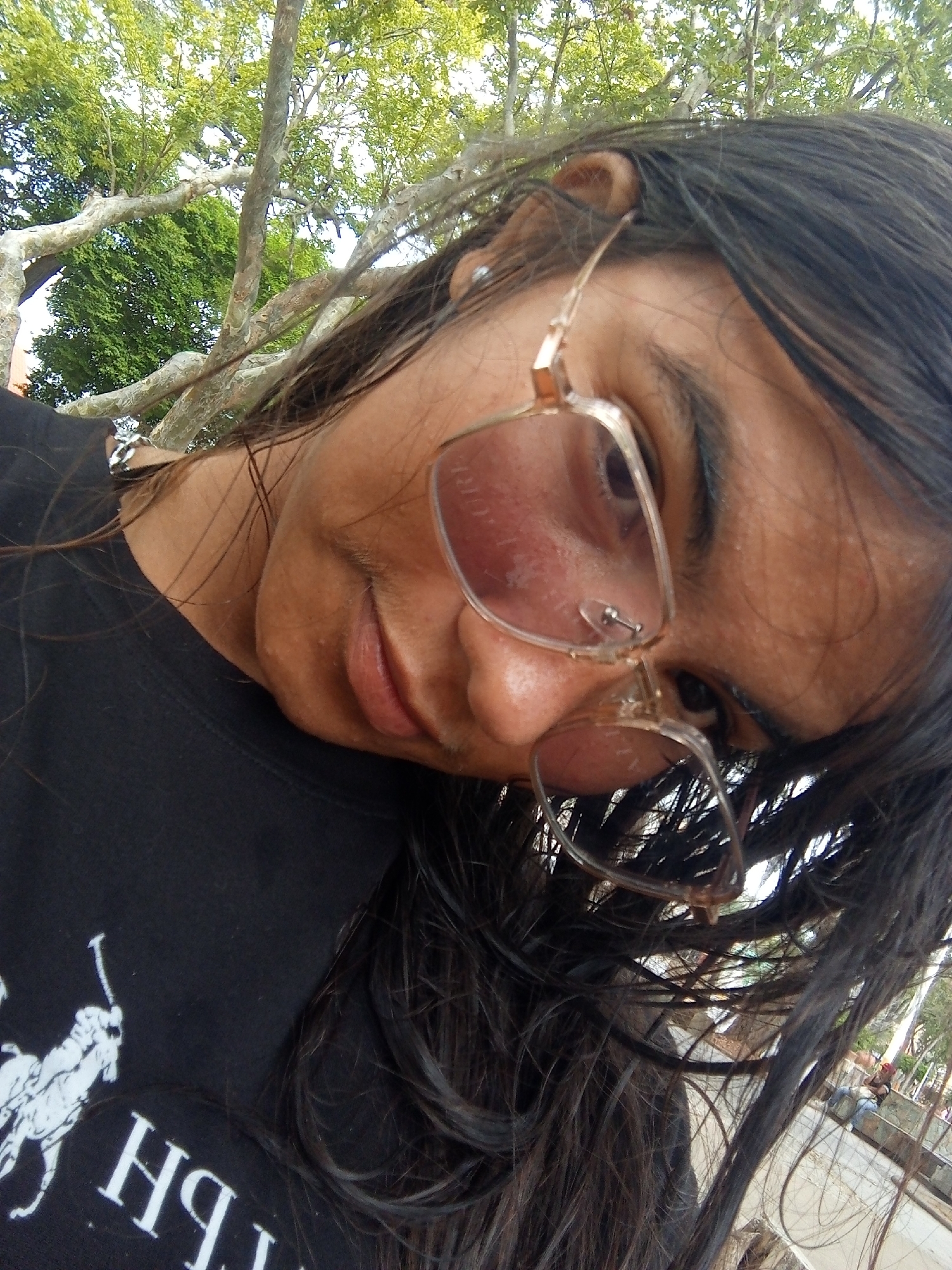 A low-angle close-up shot depicts a person with long, dark hair and tinted glasses, tilting their head towards the camera. The person is wearing a black shirt with a white logo. The background includes a tree with green foliage and a glimpse of a park area. The location is Juan Griego, Venezuela. A person sitting on a bench is visible.