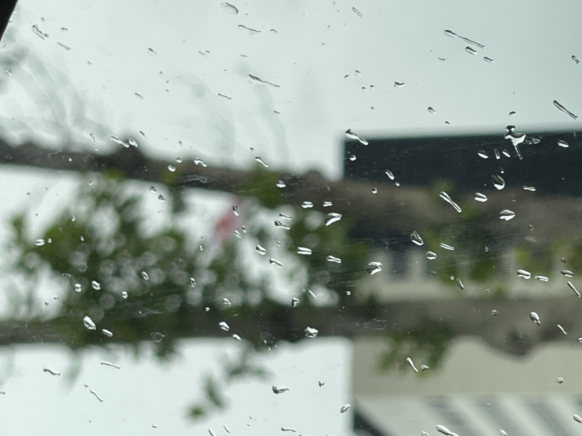 The image shows a view through a raindrop-covered window. Outside, there is a blurred background of greenery, likely trees, and a building. The building appears to be white or light-colored with a dark roof or top section. The location is potentially in the city of Tbilisi, Georgia. There is no indication of people or interactions within the frame. The focus is on the raindrops on the window and the blurred outdoor scene beyond.