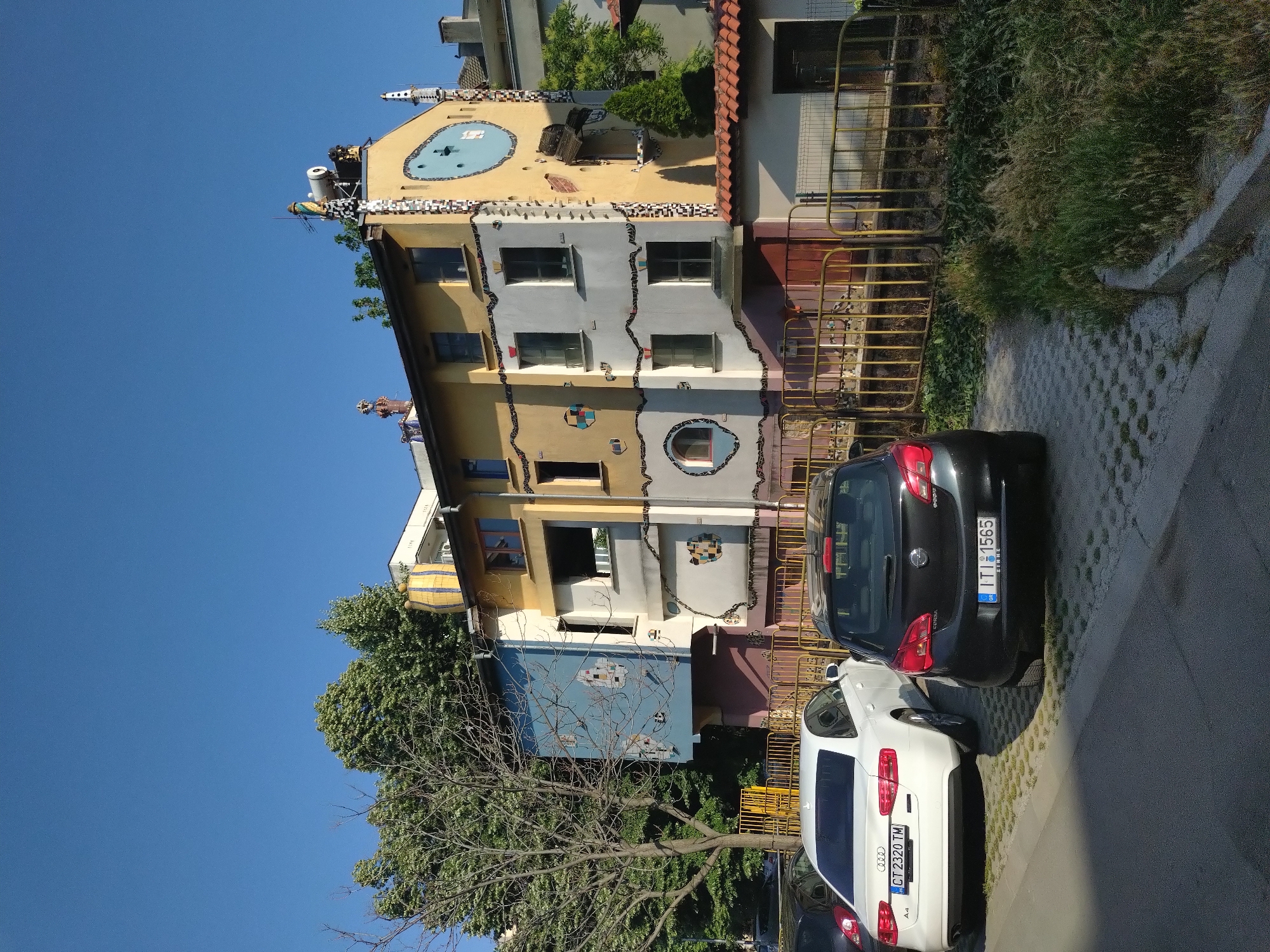 This forensic description details an outdoor, daytime scene in Stara Zagora, Bulgaria, focusing on a unique building and its immediate surroundings.
The image depicts a multi-story building characterized by an highly unconventional and artistic architectural style. The building's facade is adorned with vibrant colors, primarily yellow, white, and light blue, and extensively decorated with mosaic patterns composed of irregular ceramic or tile pieces. These mosaics form wavy lines, spirals, circles, and geometric patches across the walls. Various windows of differing shapes, including rectangular, round, and possibly oval, are irregularly placed. Sculptural elements, such as abstract forms and protruding features, are affixed to the facade. On the rightmost yellow section of the building, a large blue circular or oval design with internal markings is visible near the roofline, below which an open, broken wooden barrel-like structure extends from the wall. The roof features several unique elements, including a large, yellow and blue striped cylindrical object resembling an upturned barrel, a small figurine standing atop a chimney-like structure, a metal cylinder (possibly a water tank), and other elaborately sculpted chimney tops. A prominent light blue section of the building's facade protrudes significantly on the left side, also bearing mosaic patterns.
In the foreground, two cars are parked on a concrete paver surface with grass growing in the gaps. The car in front is a white Audi A4 sedan, with Bulgarian license plate "CT 2320 TM" (Stara Zagora). Behind it is a black Opel Corsa hatchback, bearing Greek license plate "ITI 1565 GR", with "ΣΕΡΡΕΣ" (Serres) inscribed below the numbers.
To the left of the building, a large, leafless deciduous tree with visible branches stands, while a dense canopy of green, leafy trees extends above and behind the structure. A strip of unkempt grass and weeds runs along the right side of the paved parking area, bordered by a curb. A yellow metal fence or railing separates the parking area from the building's ground level.
The sky is clear and blue, indicating a bright, sunny day. Strong sunlight casts distinct shadows, suggesting a late morning or early afternoon time. No people are visible in the scene.