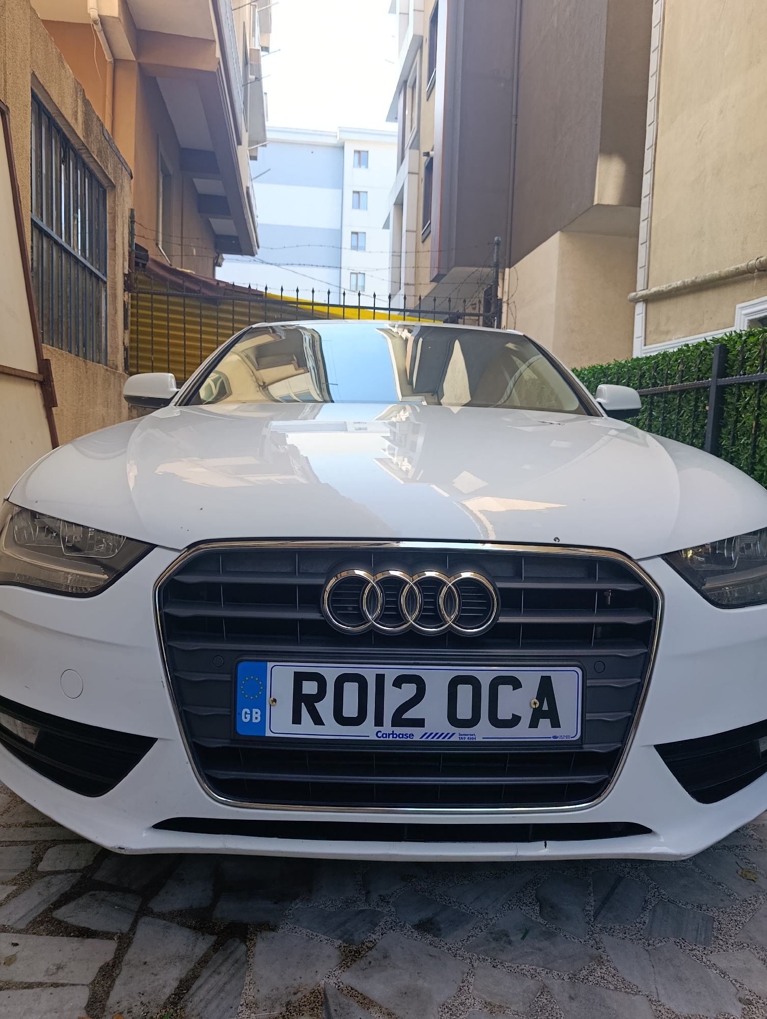A white Audi A4 sedan is parked on a stone-paved surface in front of two buildings, one with a beige facade and the other with a lighter, possibly blue and white, facade. The car's front license plate reads "RO12 OCA" and has a "GB" emblem with European stars. The car is positioned between two narrow alleyways. The right side of the image shows a dark green hedge behind a black metal fence. The left side of the image reveals a barred window and a yellow fence. The overall scene suggests an urban environment, possibly a residential area or a parking lot within a city like Ümraniye, Turkey.