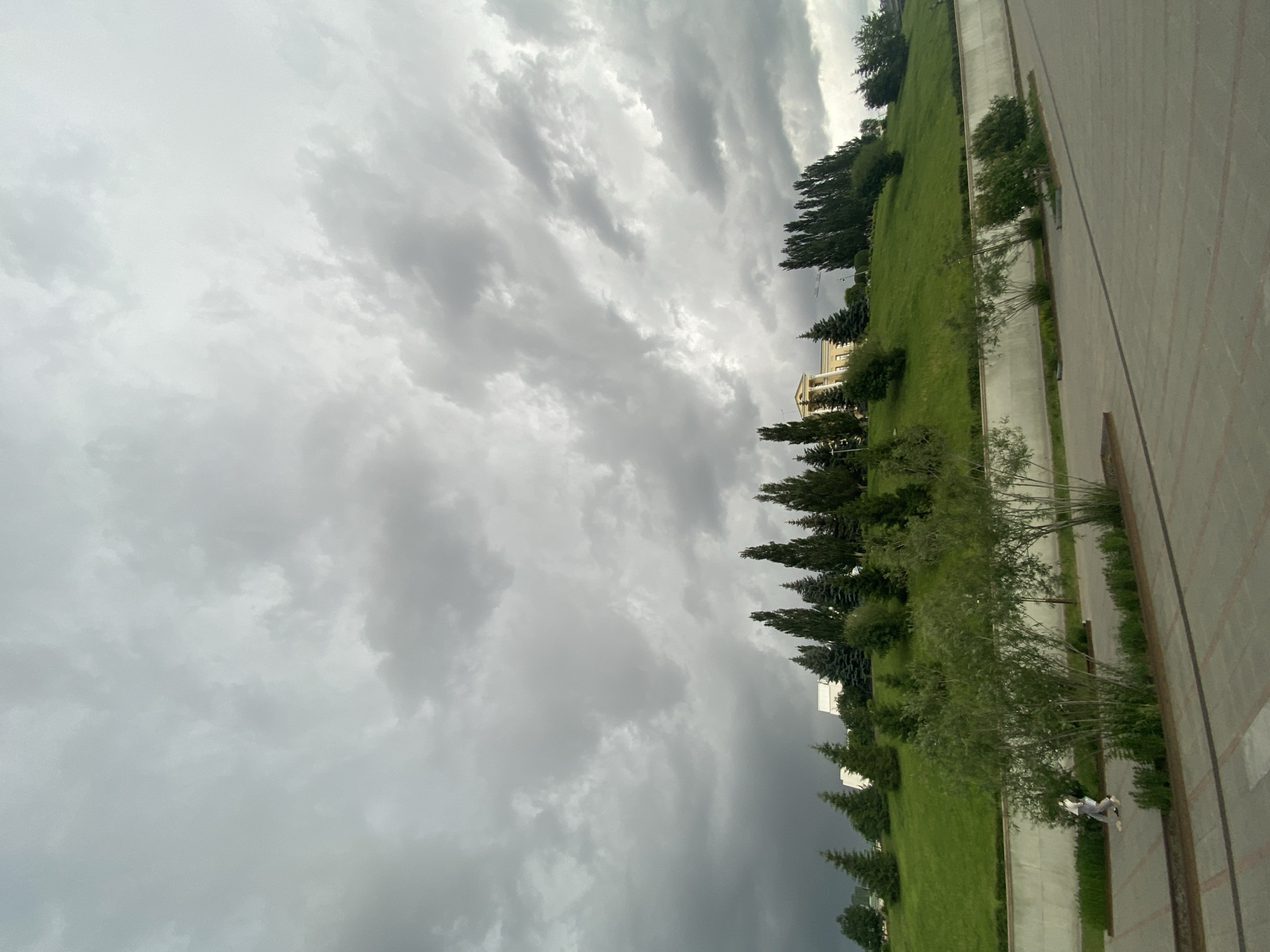 The image captures an outdoor scene dominated by a vast, heavily clouded sky, suggesting an overcast or stormy day. The sky features various shades of grey, with distinct cloud formations, some brighter and more diffused, while others appear denser and darker towards the bottom and right side of the sky.
Below the sky, the landscape consists of a wide expanse of vibrant green grass, possibly a park or large lawn, which occupies the middle left section of the frame. A line of diverse trees, including tall conifers and some deciduous varieties, runs along the right edge of this grassy area, extending into the background. Further back on the grassy expanse, a light-colored multi-story building with classical architectural details, including columns, is partially visible behind the trees. Another, less distinct white building appears further in the distance.
Separating the green space from the immediate foreground is a paved path or sidewalk that stretches horizontally across the lower part of the image. To the right of this path, there is a wider paved area, possibly a road or square, with faint markings. In the lower right corner, a single person is visible walking on the paved path. They are wearing light-colored clothing and appear to be looking down at something white held in their hands, possibly a book or papers.
The time of day is daytime, but the heavy cloud cover creates diffused light and a somewhat somber atmosphere. There is no visible text in the image. The setting appears to be an urban park or public green space.