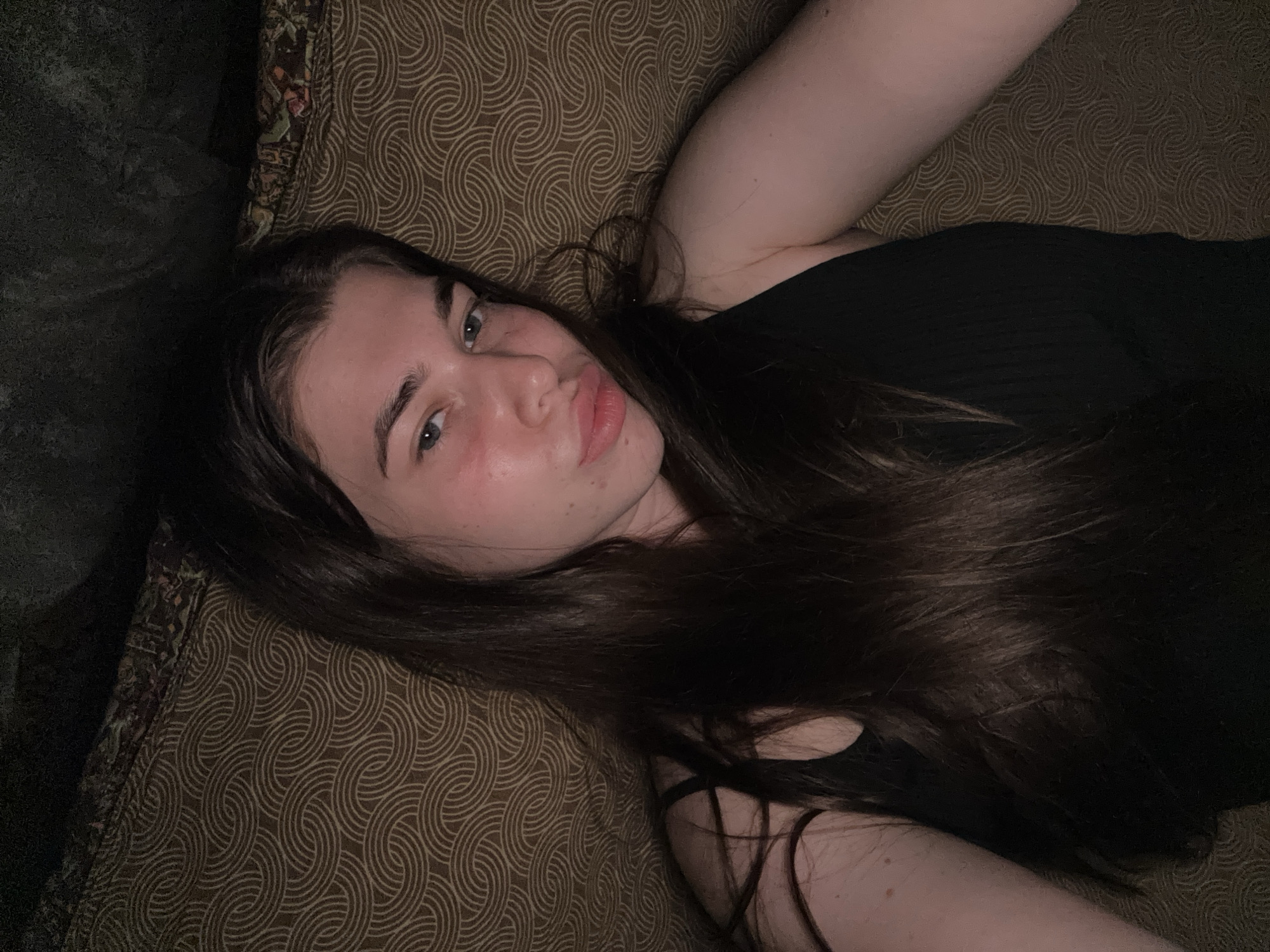 A young woman with long, dark brown hair is lying down, looking towards the camera with a pouty expression. She is wearing a black tank top. Her face is lightly flushed, and she has some freckles on her nose and cheeks. She is lying on a patterned surface that appears to be a bed or sofa. The background is dark and out of focus, suggesting an indoor setting. The image does not provide any specific clues to indicate the location in the city of Мещёры, Russia.