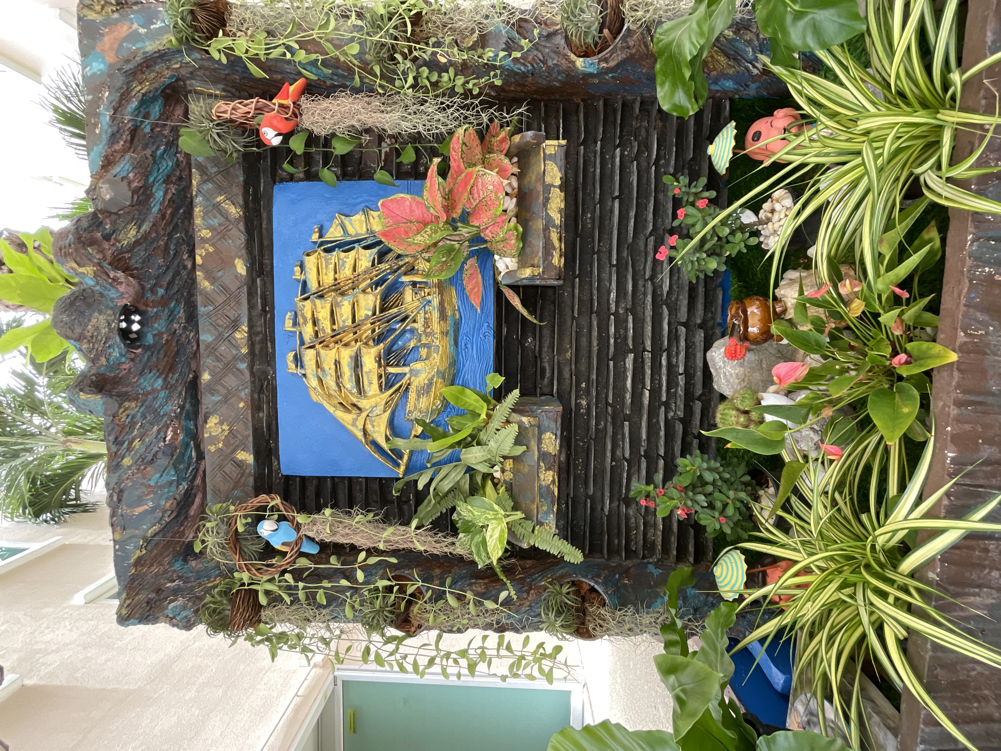 The image captures an outdoor decorative fountain, likely situated in a garden or public space within Pattaya City, Thailand. The fountain features a dark, textured backdrop with blue and gold accents, resembling rock formations. A relief of a golden galleon on a blue sea is mounted centrally. Various plants, including ferns and trailing vines, are incorporated into the fountain's design, some hanging in woven baskets. Two ceramic parrot figurines, one blue and one red, are perched on the fountain. The lower section of the fountain contains a variety of potted plants, including a spiky, variegated green and yellow plant in the foreground, and flowering plants with pink and red blooms. Small decorative elements, such as a miniature umbrella, a pink anthurium flower, and a smiling pink figurine, are visible among the plants. The background includes a portion of a building with cream-colored textured walls and a palm tree frond. The overall scene is well-lit, suggesting it was captured during daylight.