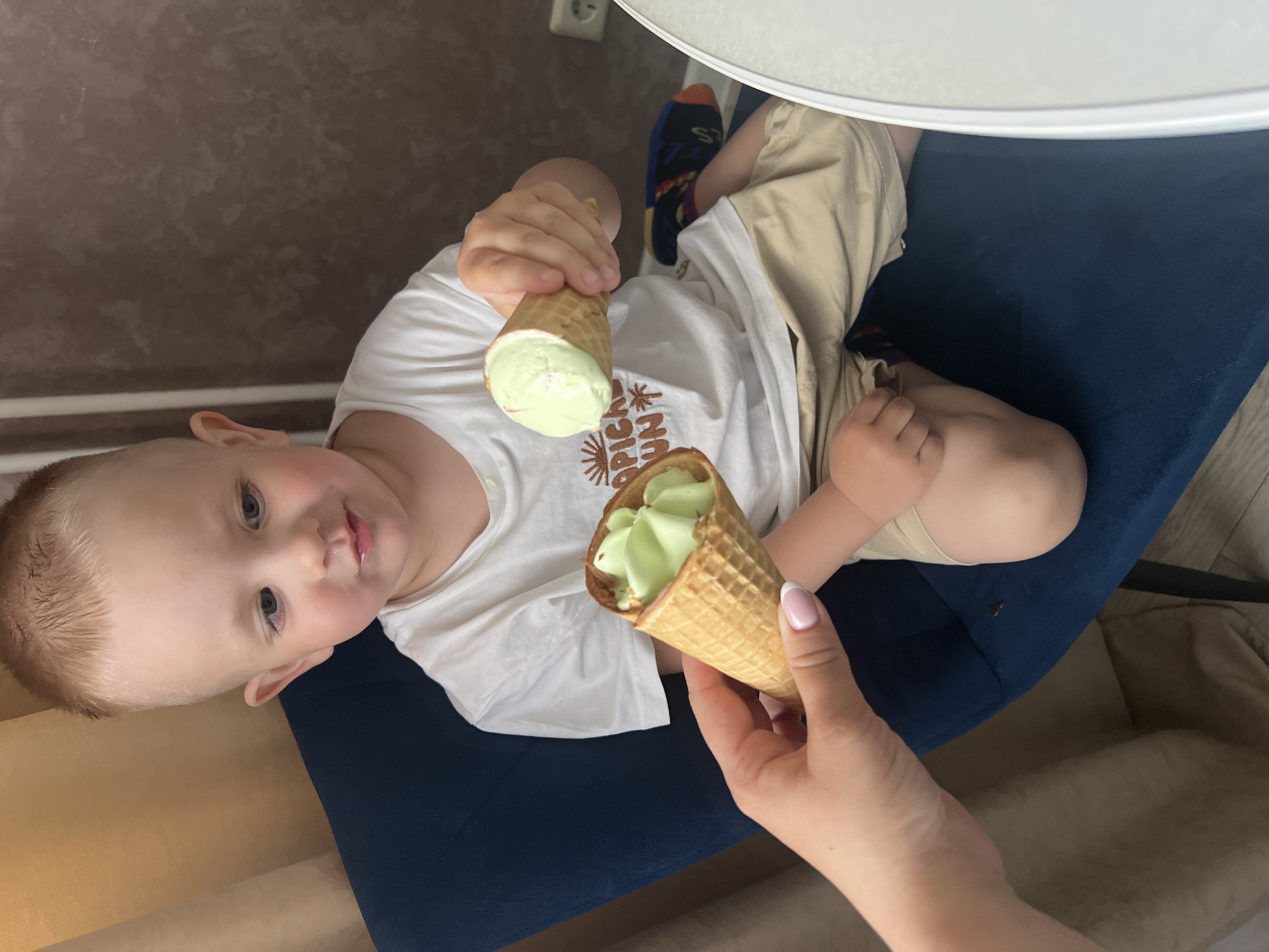 The image, rotated 90 degrees clockwise, captures an indoor scene featuring a young child and an adult's hand sharing ice cream. A fair-skinned toddler with light hair is positioned on a dark blue upholstered surface, likely a sofa or chair cushion. The child looks directly towards the camera with a neutral, slightly curious expression.
The child is wearing a white t-shirt, which has a brown, stylized graphic on the front partially visible, including the text "TROPICAL SUN." They also wear beige shorts and colorful socks with blue, orange, and black patterns. The child holds a waffle cone filled with light green ice cream in their left hand. An adult's hand, with manicured nails featuring French tips, extends into the frame from the right, holding a second identical waffle cone also filled with the same light green ice cream, suggesting an act of offering or sharing.
The background includes a textured brown wall on the left side of the frame, where a white electrical outlet is visible. A bright white surface, possibly a table or countertop, is partially visible at the top. The overall setting is a casual, domestic environment, likely in Марковский, Russia. The scene conveys a simple, pleasant moment of enjoyment.