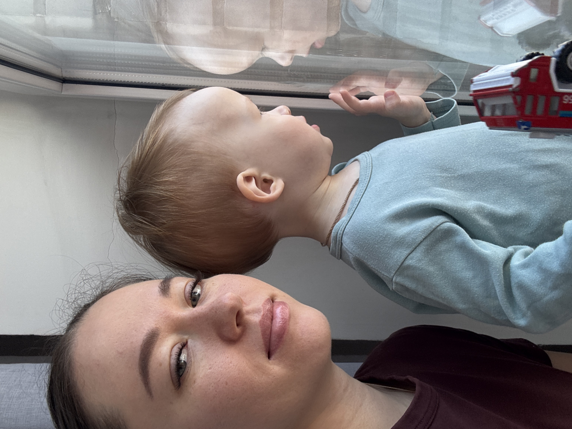 A woman and a child are lying down inside a building, possibly Ferma, Russia. The child is looking up at a window with his hand placed on the glass. A toy truck rests near the child on their clothing. The woman is lying close to the child and looking at the camera.