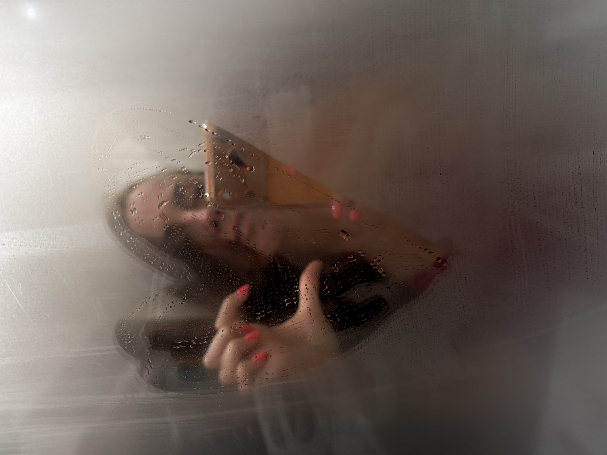 The image captures a close-up view of a person seen through a steamy, water-streaked glass or plastic surface. The setting is indoors, likely a bathroom or shower enclosure in Birobidzhan, Russia, given the humid environment suggested by the condensation.
The primary subject is a person, whose face and hands are pressed against or are very close to the translucent surface. The person appears to be a woman, with long dark hair and hands featuring bright red nail polish. Her face is partially obscured by the steam and water droplets, making her features indistinct, but a slight smile is discernible. Her head is tilted, and her eyes seem to be looking outwards or downwards.
One hand is prominently displayed in the lower-left portion of the frame, with fingers spread and resting against the surface, showcasing the vibrant red nails. Another hand is visible further to the right, also with painted nails, though less distinct. The surface itself is covered with varying densities of steam and numerous water droplets, some running down in streaks, creating a soft-focus, ethereal effect.
The background behind the person is a blurred, indistinct area of lighter and darker tones, suggesting a wall or interior space. No specific objects or further details of the background are clearly visible due to the heavy obfuscation. The activity depicts an interaction with the steamy surface, possibly a moment of playfulness, relaxation, or introspection. There is no visible text in the image.