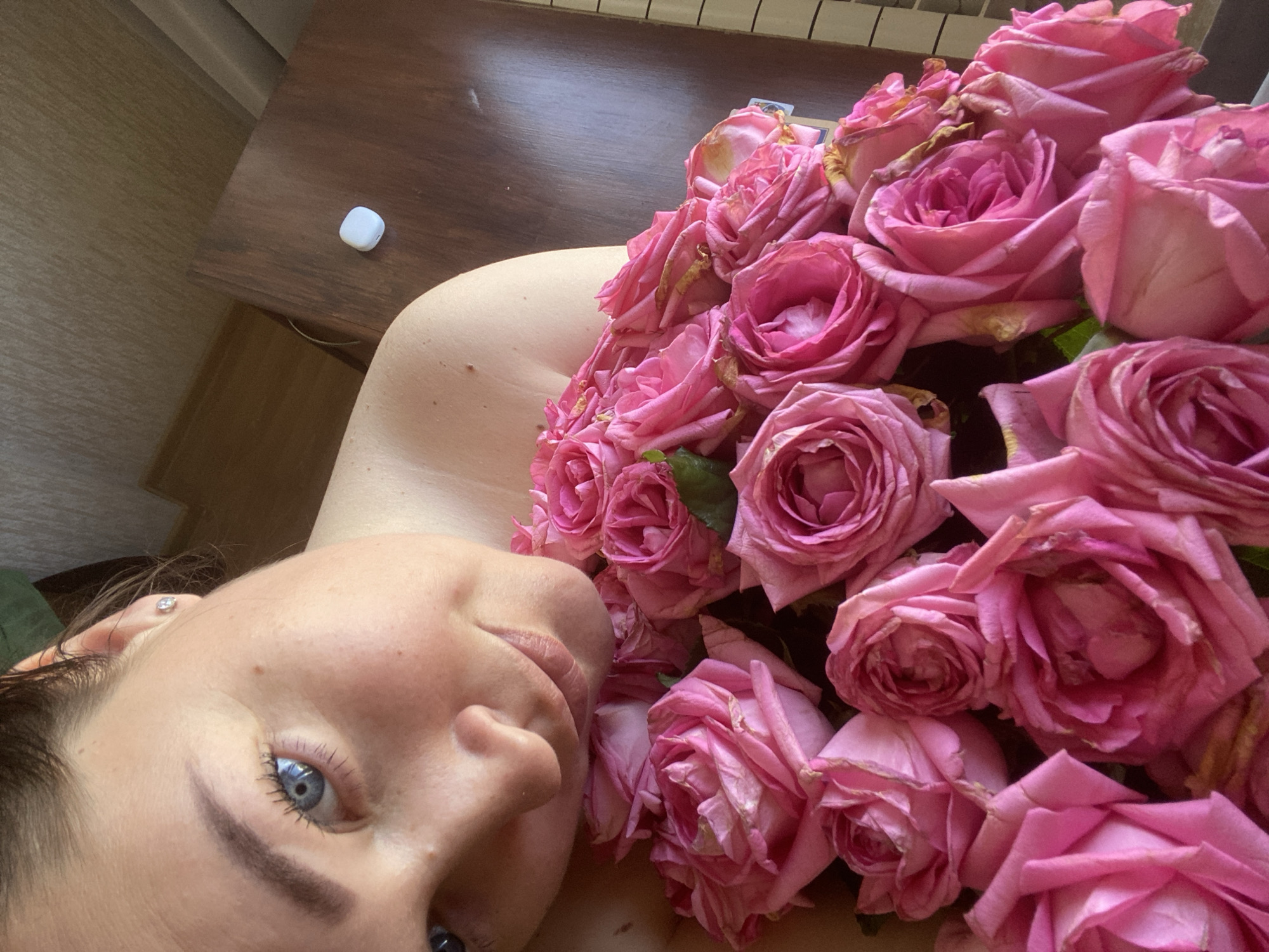 A fair-skinned woman with blue eyes is lying down, partially covered by a large bouquet of pink roses. A portion of a wooden table with a small, white, rectangular object is visible in the background. The location of the scene is unknown; there is nothing in the image to suggest that it is in Sochi, Russia.