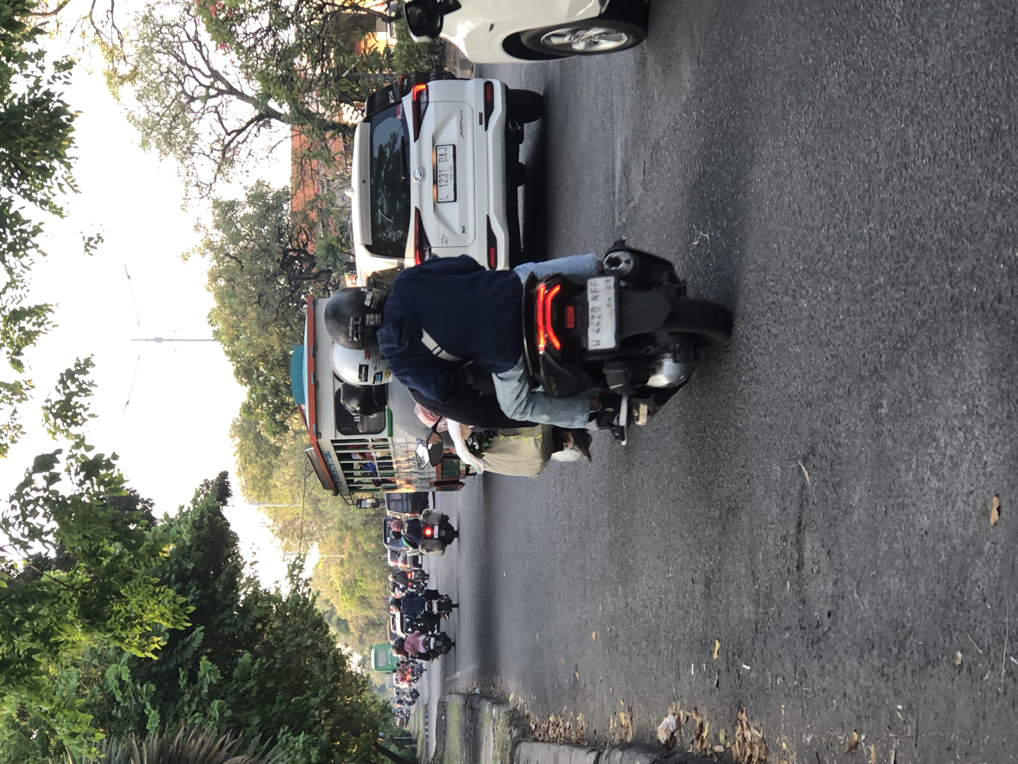 The image captures a busy street scene in Ngampelsari, Indonesia, likely during the late afternoon or early evening, given the lighting. The primary focus is a person riding a motorcycle in the foreground, seen from behind, wearing a dark blue hoodie and light-colored jeans. They are on a dark-colored motorcycle, which has a license plate showing "W 4420 NFF". To their right, a white SUV is partially visible, with a license plate reading "L 1231 DAJ". In the background, a long, multi-passenger bus with an open-sided design is driving on the road, filled with passengers. Numerous other motorcycles and some cars are also present, indicating moderate traffic. The road is paved, with a curb and foliage lining the left side. Trees with green leaves overhang the street, suggesting a semi-urban or suburban environment. The overall impression is one of a typical day on Indonesian roads, with a variety of vehicles and pedestrians navigating the traffic.