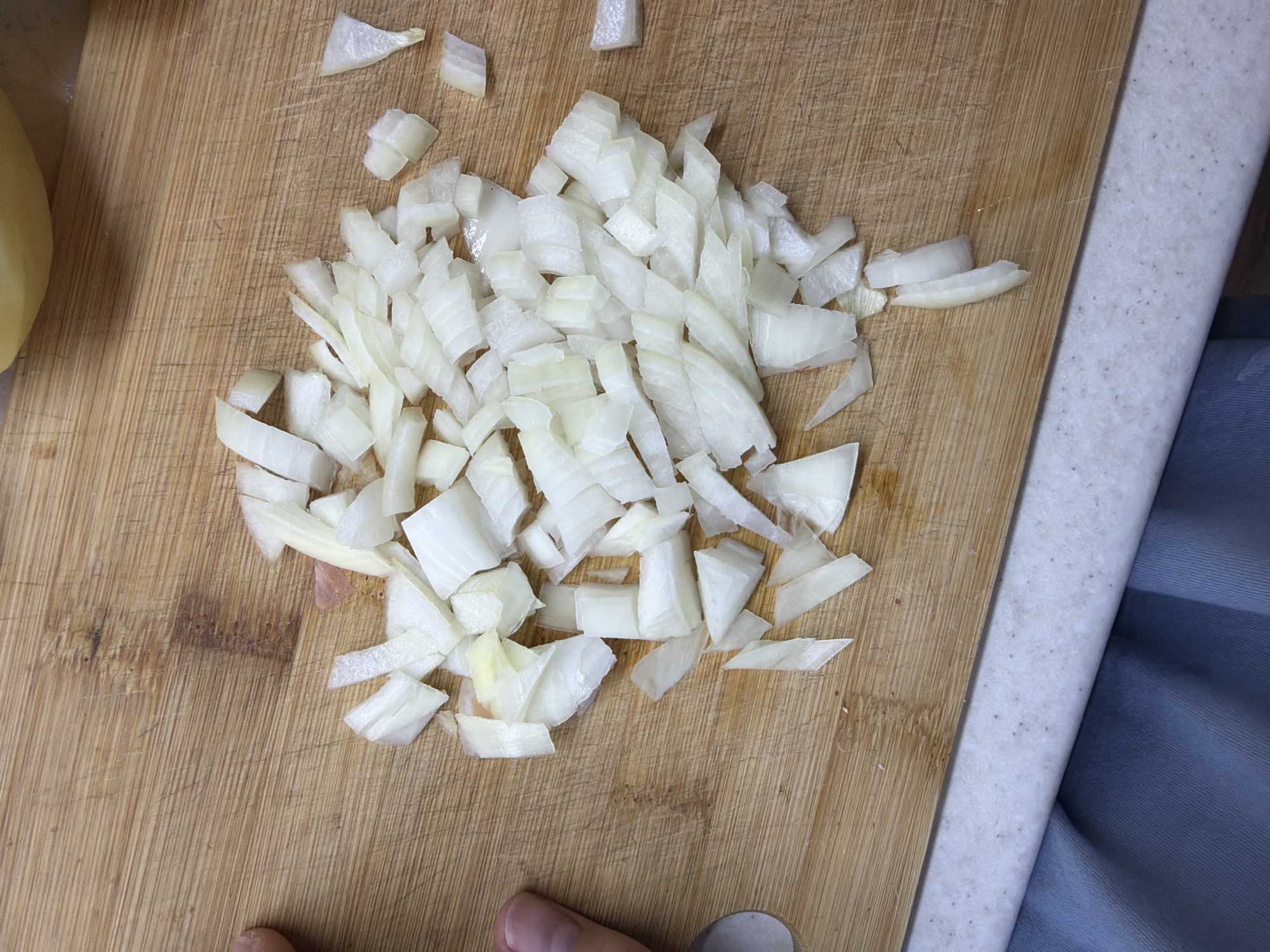 The image presents an overhead, close-up view of a kitchen scene, located in Chaikovsky, Russia, focused on food preparation. A substantial pile of roughly chopped white onion pieces occupies the central area of a light brown wooden cutting board. The onion pieces vary in size and shape, generally appearing white with some segments exhibiting a faint yellowish tint, and some moisture is visible on the board. The cutting board itself displays numerous parallel knife marks, scratches, and a few faint reddish-brown stains, indicative of prior use. In the upper right corner, a curved, light yellow object, likely a peeled potato, is partially visible and out of focus. A human left thumb and the adjacent part of a forefinger are visible on the cutting board along the left edge, positioned near a sliver of a silver-colored utensil or knife handle. The cutting board rests on a speckled white countertop, which is visible across the bottom portion of the frame, with a soft, draped segment of blue or grey fabric visible below the countertop.