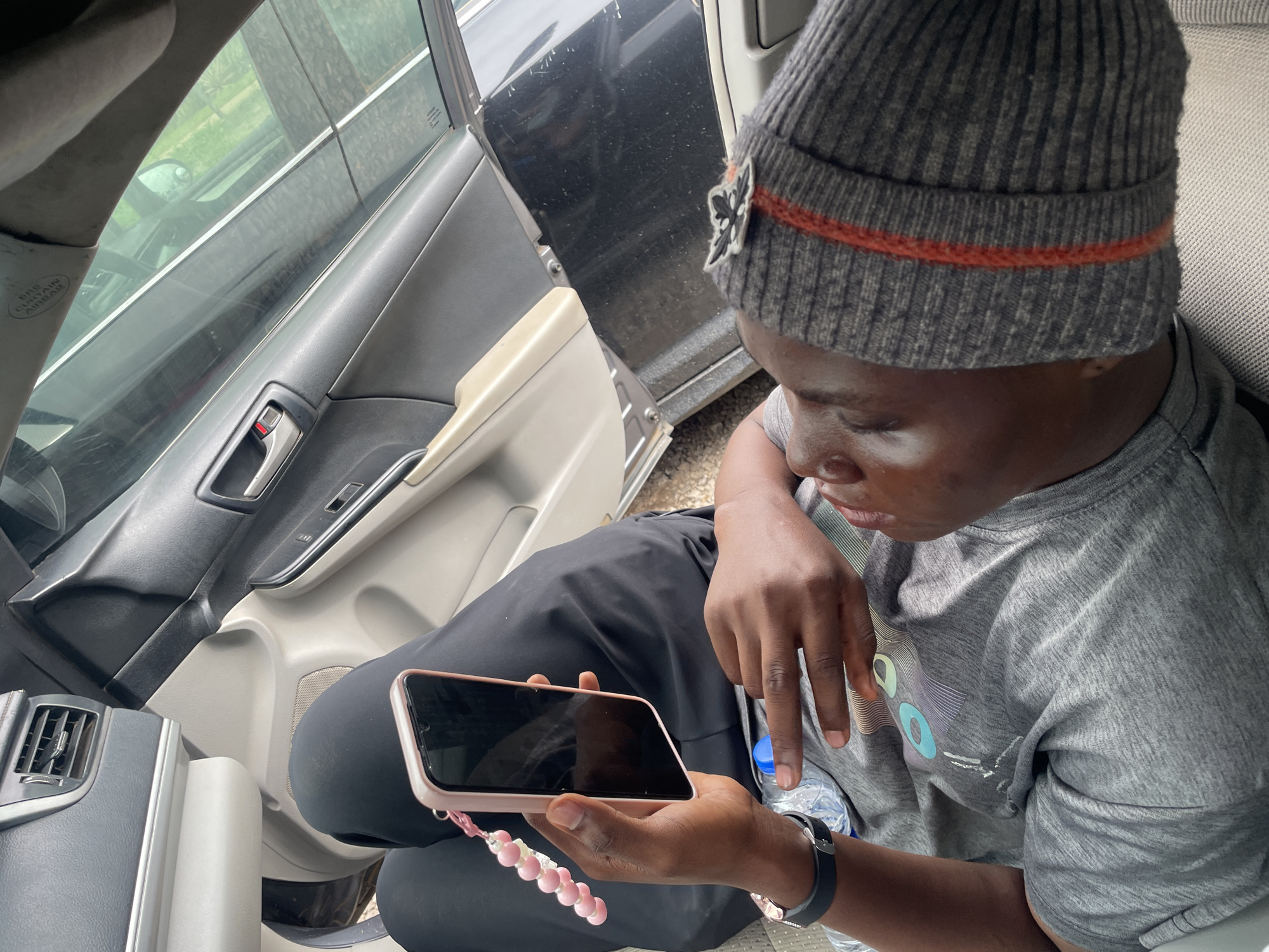A person is sitting in a car in Galadimawa Village, Nigeria, looking at their phone. The person is wearing a grey t-shirt, black pants, and a grey beanie with an orange stripe. They are holding a pink phone with a beaded charm attached. A water bottle is visible near their hands. The interior of the car, including the dashboard, door panel, and a vent, is visible. Sunlight is streaming through the car's windows.