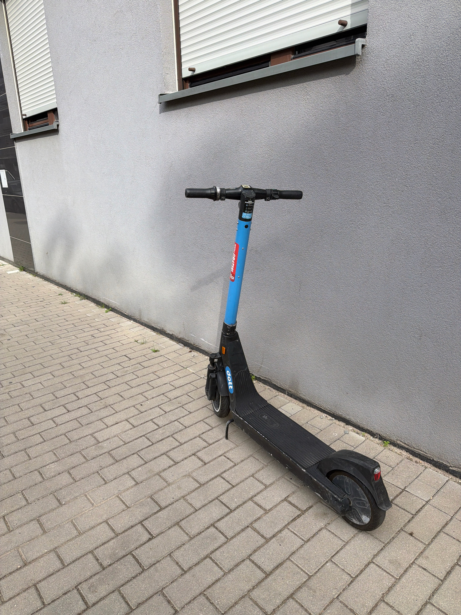 A blue and black electric scooter, branded "Dott", stands on a brick sidewalk next to a textured grey building. The building has a window with a closed white rolling shutter and a metal ledge. The scooter is upright and appears to be parked. The sidewalk is paved with rectangular bricks arranged in a staggered pattern. There is some sparse vegetation growing along the base of the building. The scene is illuminated by daylight. The image was taken in Wrocław, Poland.