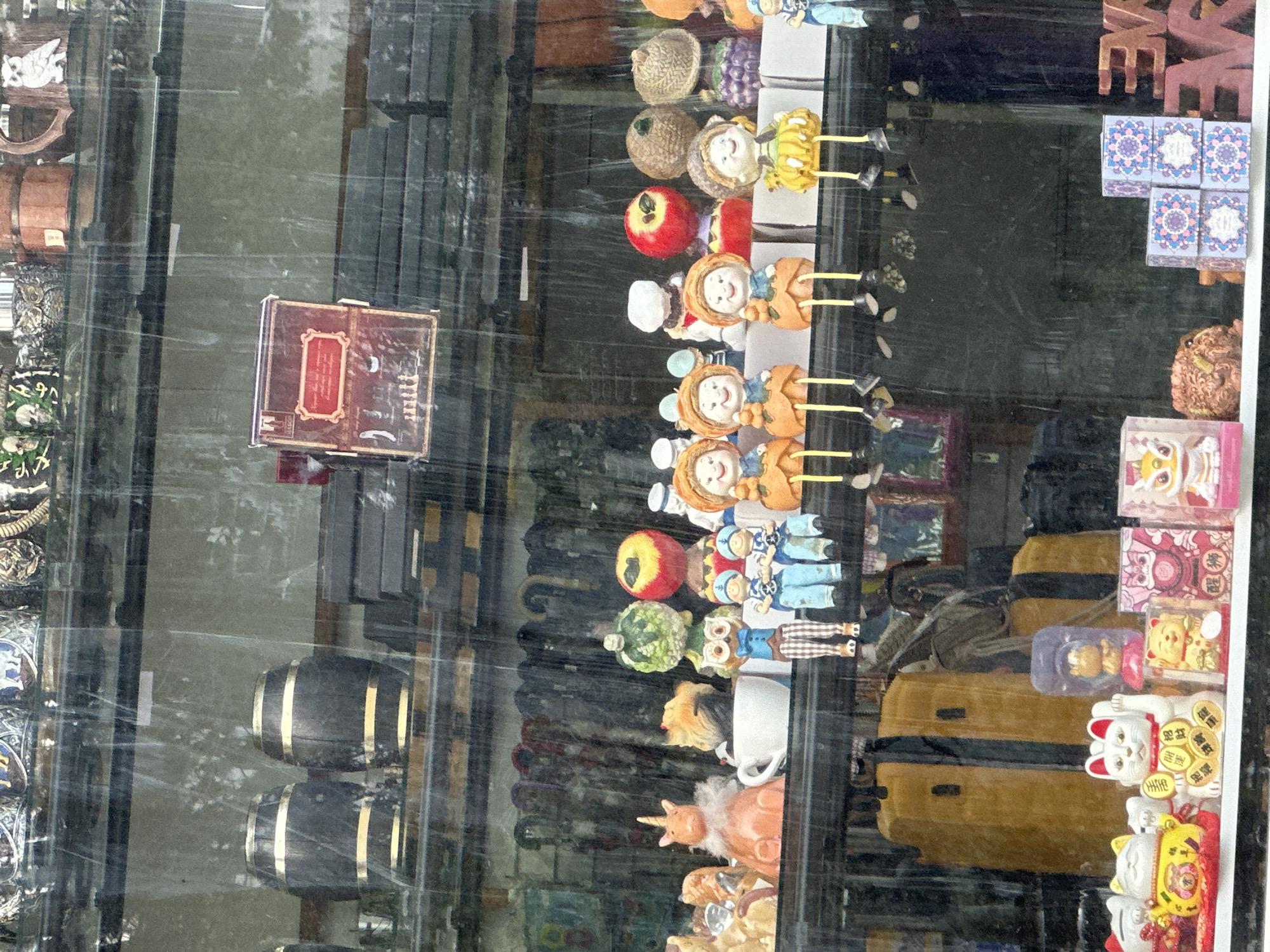 This image shows a street-level view of a shop window, with raindrops on the glass distorting the view of the merchandise. The items on display are diverse, including decorative figurines, ceramic cats, boxes with intricate patterns, and what appear to be two wooden barrels on an upper shelf. The overall impression is of a cluttered shop selling various curios and souvenirs. It is not possible to determine the exact location in Sochi, Russia, or any specific actions or interactions.