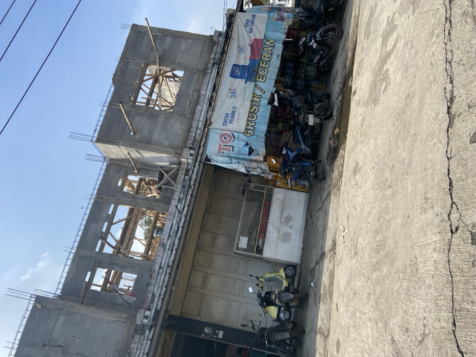 The image shows a street scene in Margaasih, Indonesia (latitude: -6.942, longitude: 107.551). There is a partially constructed building with exposed wooden framing and concrete blocks. In front of the building, several motorcycles are parked. A shop selling groceries and other items is located at the bottom right of the image. The shop sign reads "TOKO" (Indonesian for "store") and "GROSIR / ECERAN" (Indonesian for "wholesale/retail"). The street is paved concrete and there is a small puddle of water in front of the shop. The sky is mostly clear, with a few wispy clouds. A building across the street is visible in the background. No people are visible.
