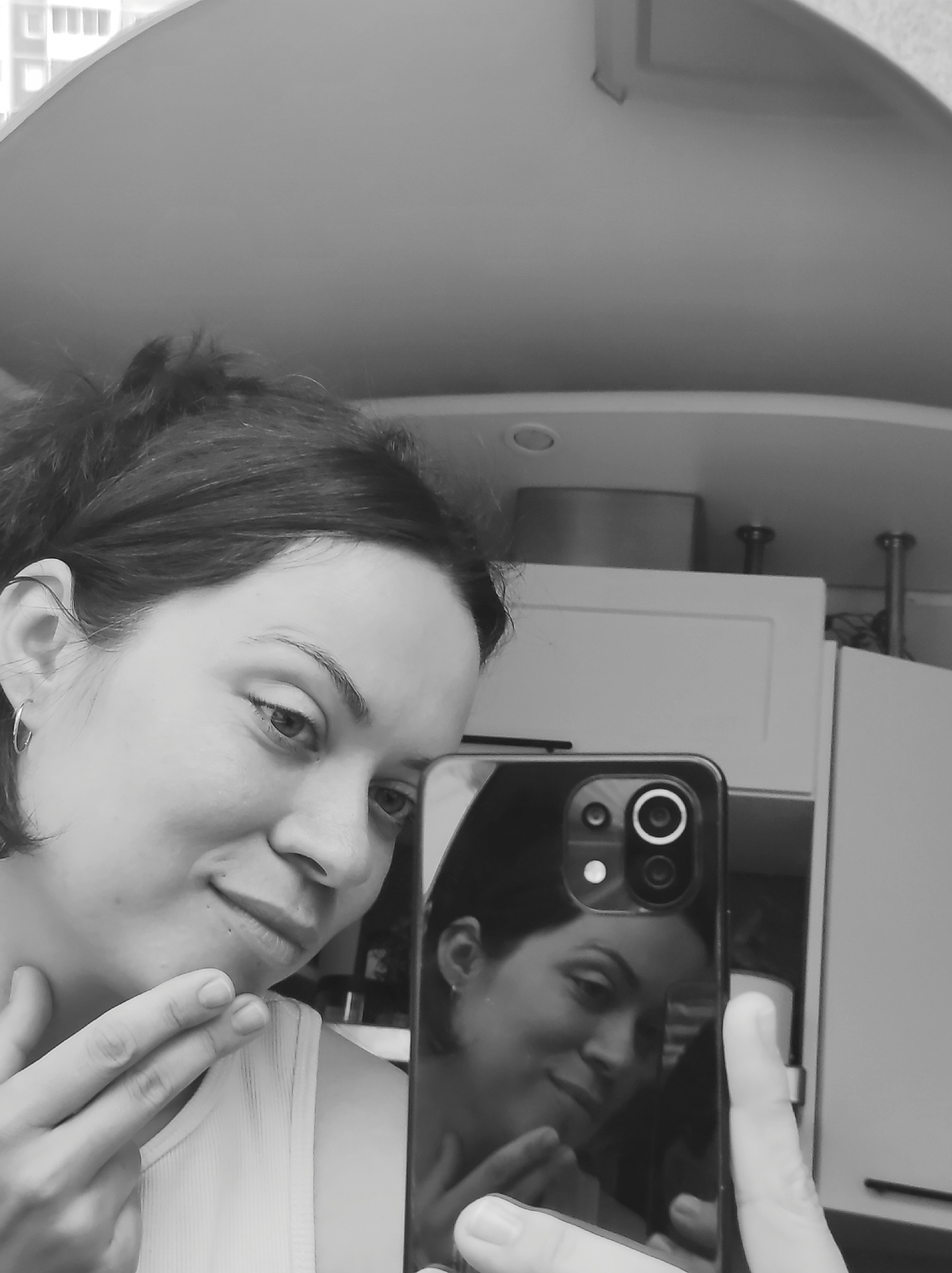 A woman takes a selfie in a kitchen, possibly in Chaikovsky, Russia. The woman has dark hair pulled back, and she is wearing a tank top. She holds a smartphone in her right hand, using it to capture her reflection. Her left hand is near her chin. The background reveals kitchen cabinets and a curved ceiling. The image is in black and white.