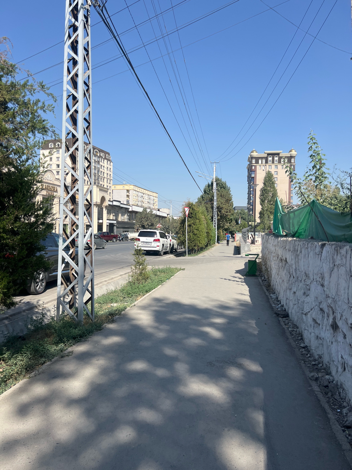 The image depicts a street scene in Osh City, Kyrgyzstan, under a clear blue sky. A paved sidewalk runs along the right side of the image, bordered by a rough, light-colored stone wall and then a green tarp on top of some construction or landscaping. The sidewalk is partially shaded by the shadows of trees. To the left of the sidewalk is a street with parked and moving vehicles. In the foreground on the left, a tall metal utility pole with a lattice structure is prominent, with power lines extending overhead across the sky. Behind the pole, lush green trees and bushes are visible. In the background, several multi-story buildings line the street, some with ornate architectural details and arched windows. One tall building on the right has a distinctive brown and beige facade with gold-domed accents at the top. Several cars are on the street, including a white SUV and a dark-colored sedan parked near the utility pole. A few people are visible in the distance, one walking along the sidewalk and others near the buildings. The environment appears to be an urban setting with a mix of residential and commercial buildings, trees, and infrastructure.