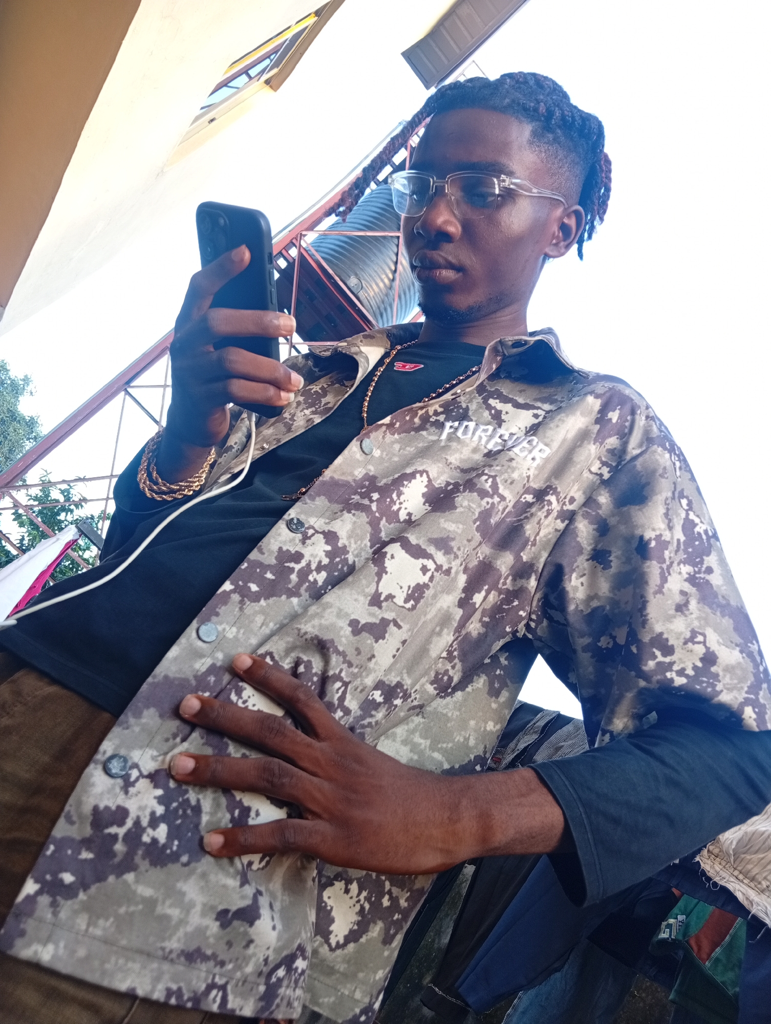 A young Black man, wearing glasses, a black t-shirt, a camouflage jacket with "FOREVER" printed in white on the sleeve, and brown pants, holds a black smartphone in his left hand. He is also wearing a gold chain and a gold bracelet on his left wrist. His right hand is placed on the front of his jacket. The man appears to be looking down at his phone. The background suggests an outdoor setting with a bright sky, some metallic structures, and hints of greenery. The lighting indicates it is daytime, likely sunny. The scene appears to be taking place in Ukukwa, Nigeria.