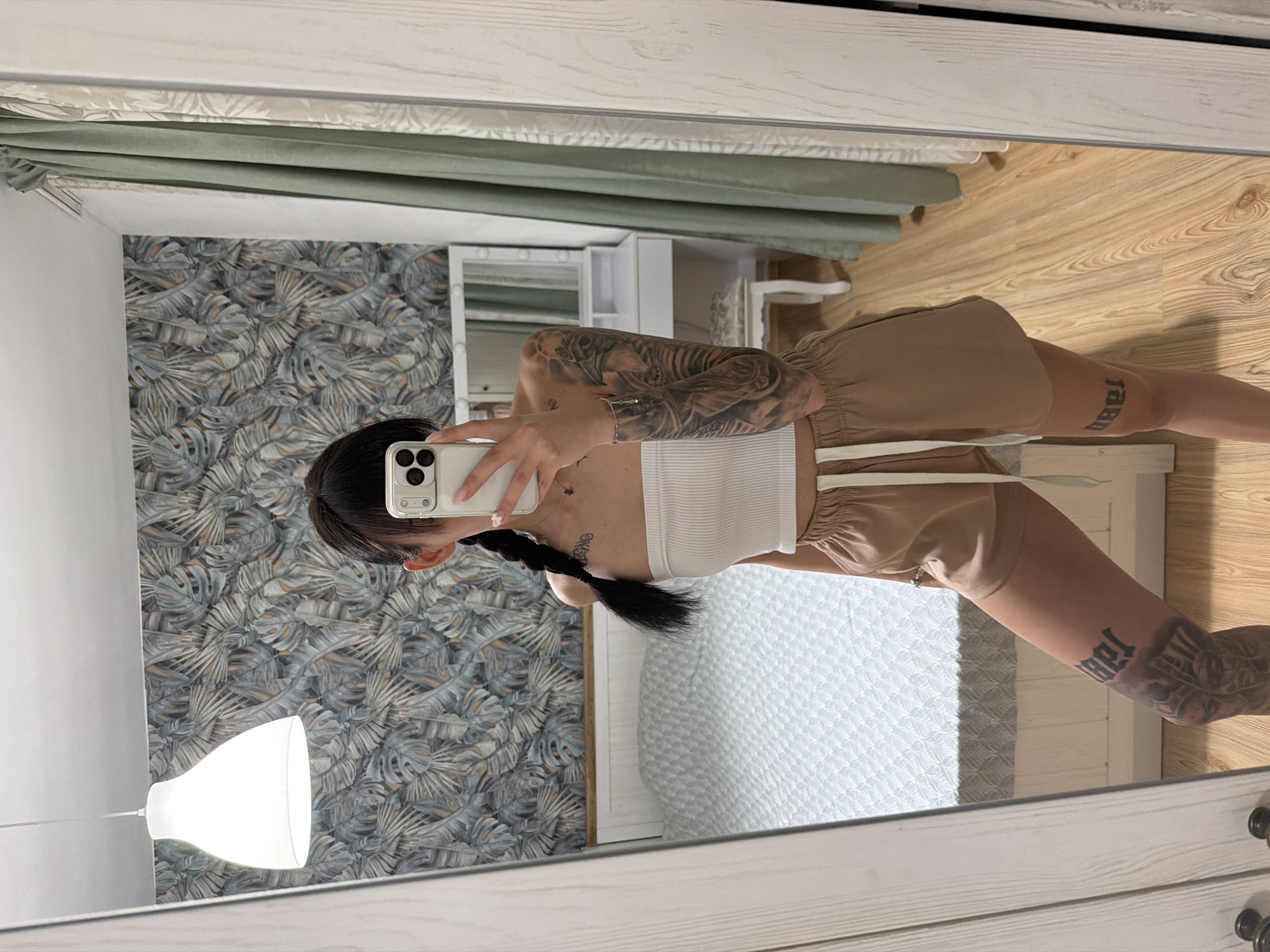 A young woman with dark hair in a braid is standing in front of a mirror, taking a selfie. She is wearing a white strapless top and tan shorts. The woman has tattoos on her arms and legs. The room appears to be a bedroom, with a bed in the background and patterned wallpaper on the wall. The image was taken in Plovdiv, Bulgaria.